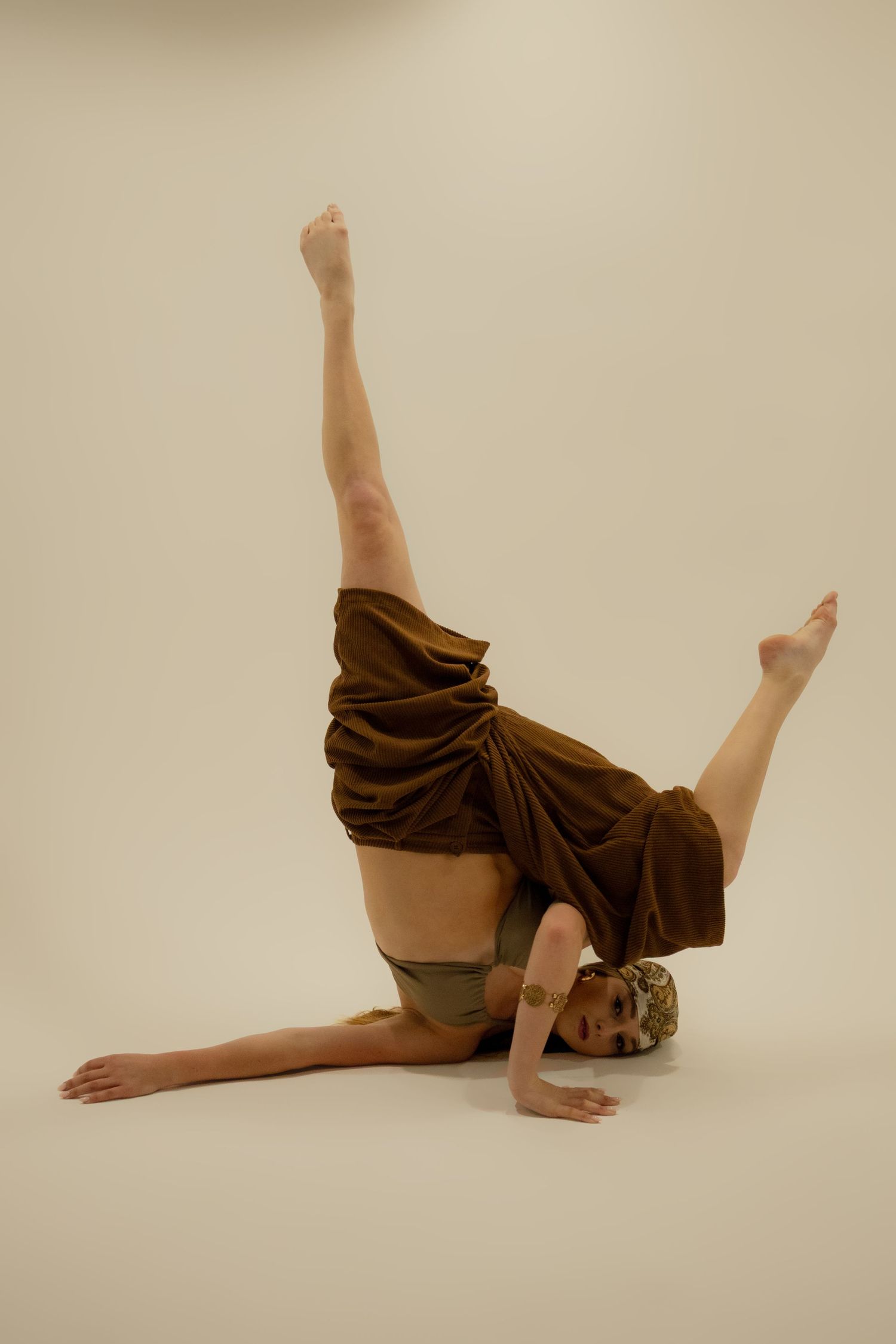 Dancer performing advanced floor pose with legs raised in brown draped costume.