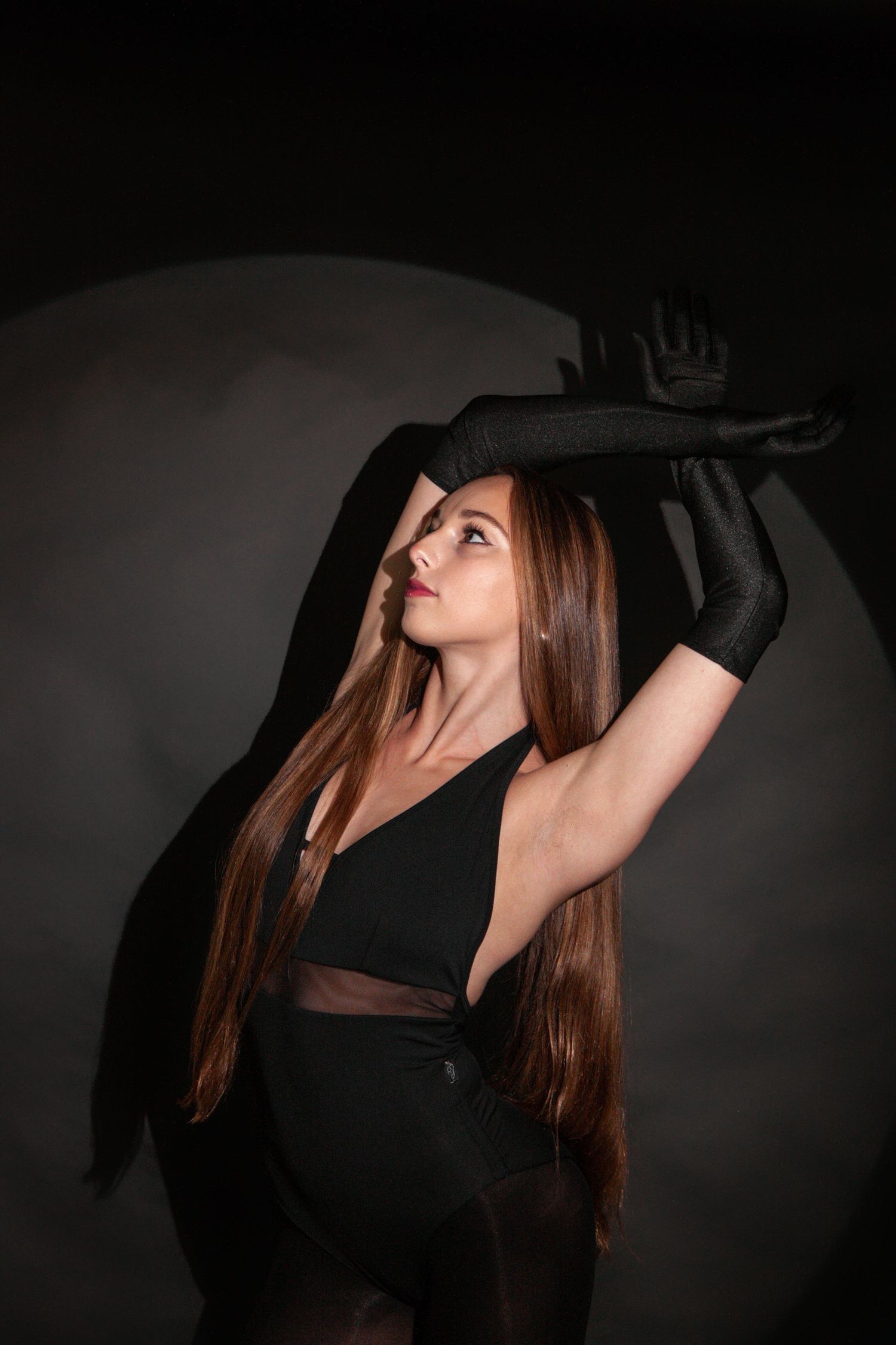 Dancer in black bodysuit and gloves strikes dramatic pose with raised arm in dark studio.