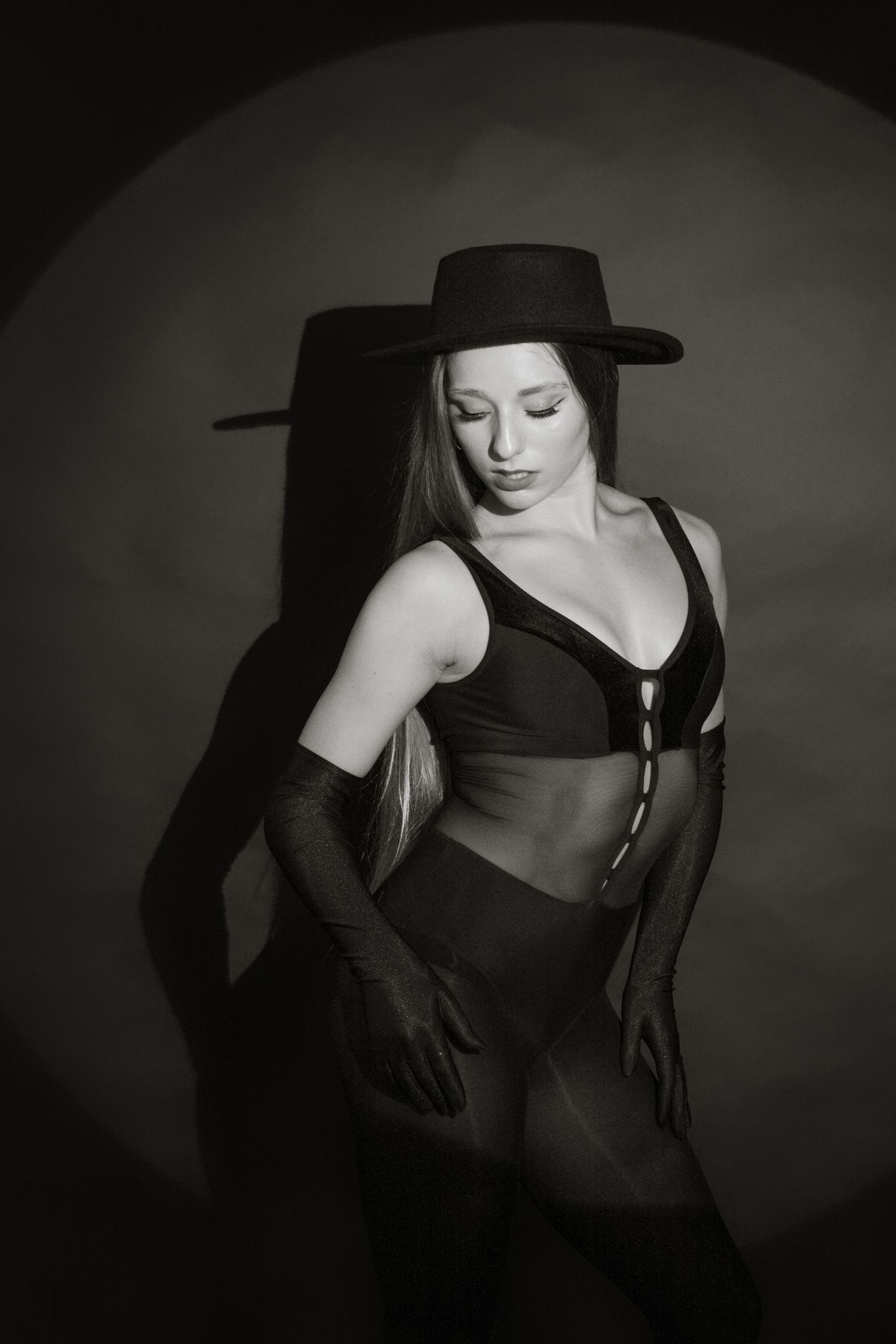Black and white portrait of woman in bodysuit and wide-brim hat posing in moody spotlight studio setting.