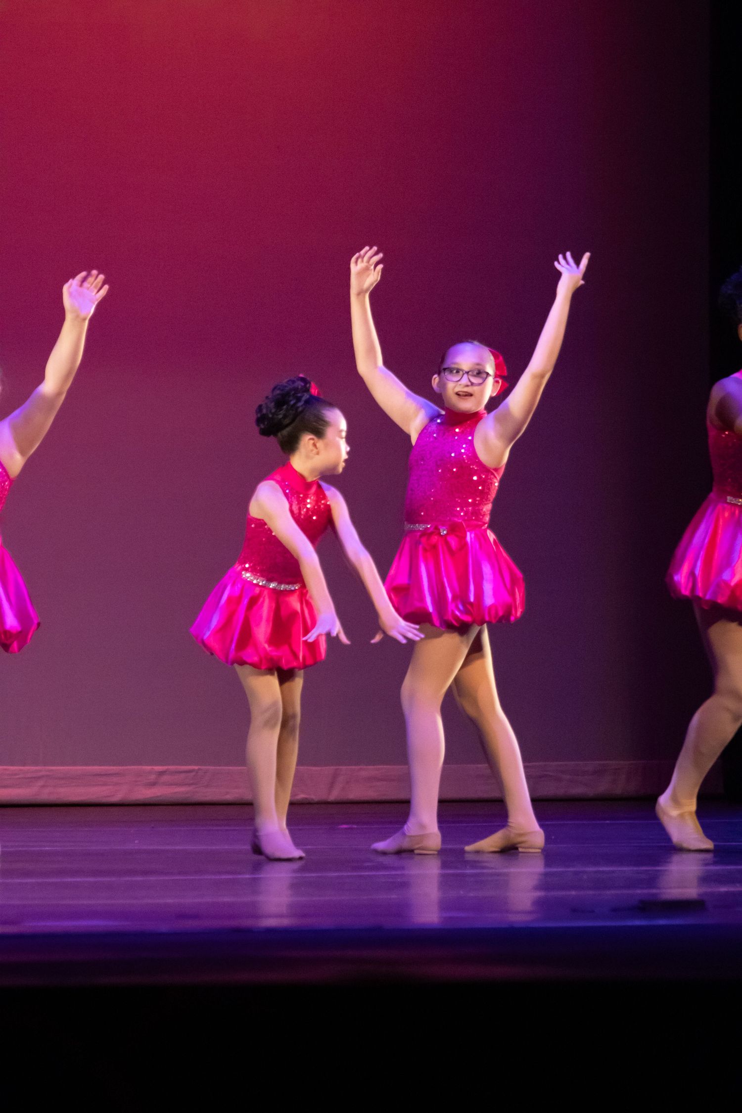 Young girls in pink sequined costumes perform a dance recital on stage under warm spotlight lighting.