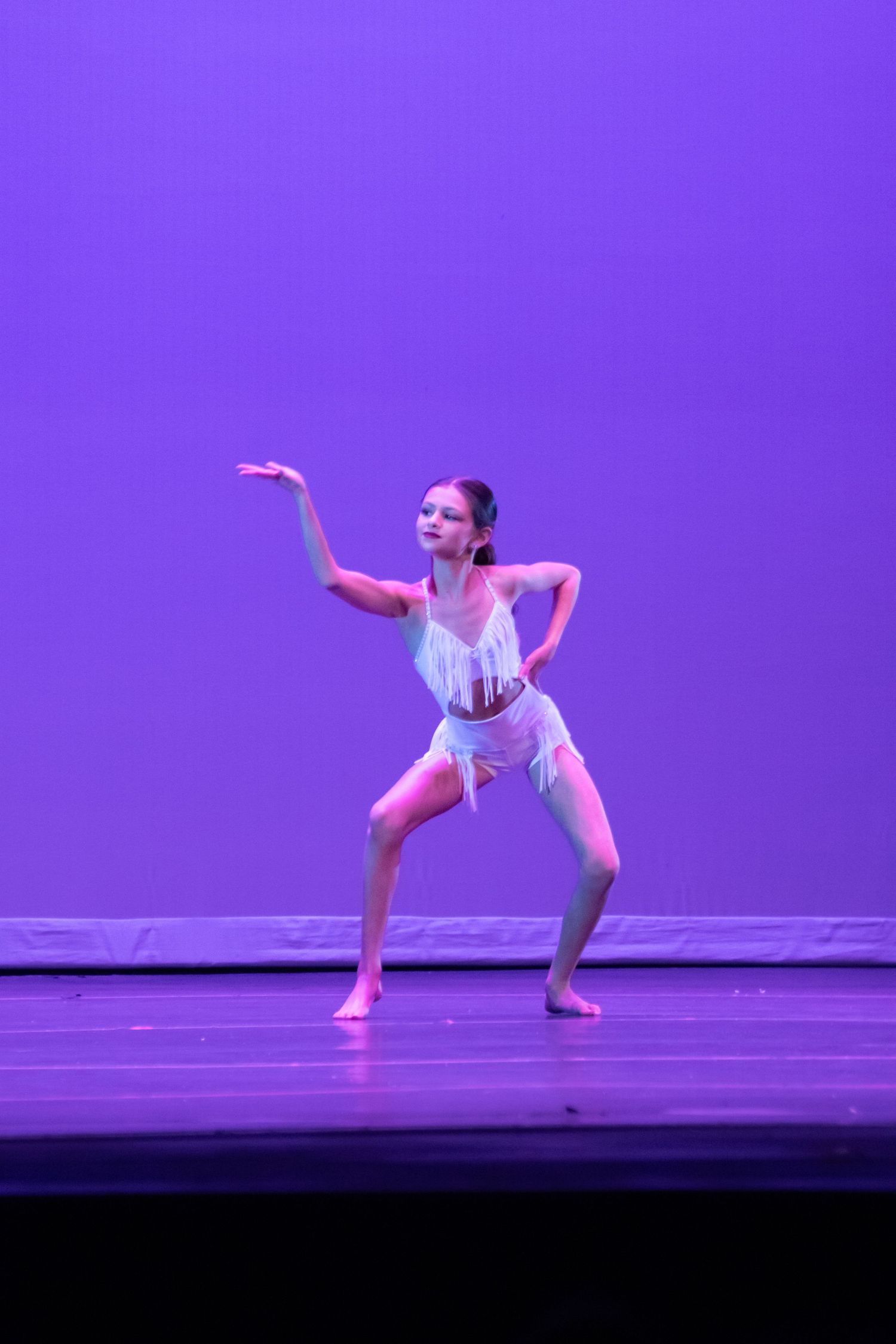 A young girl performs a contemporary dance solo in a white fringe costume under purple stage lighting.