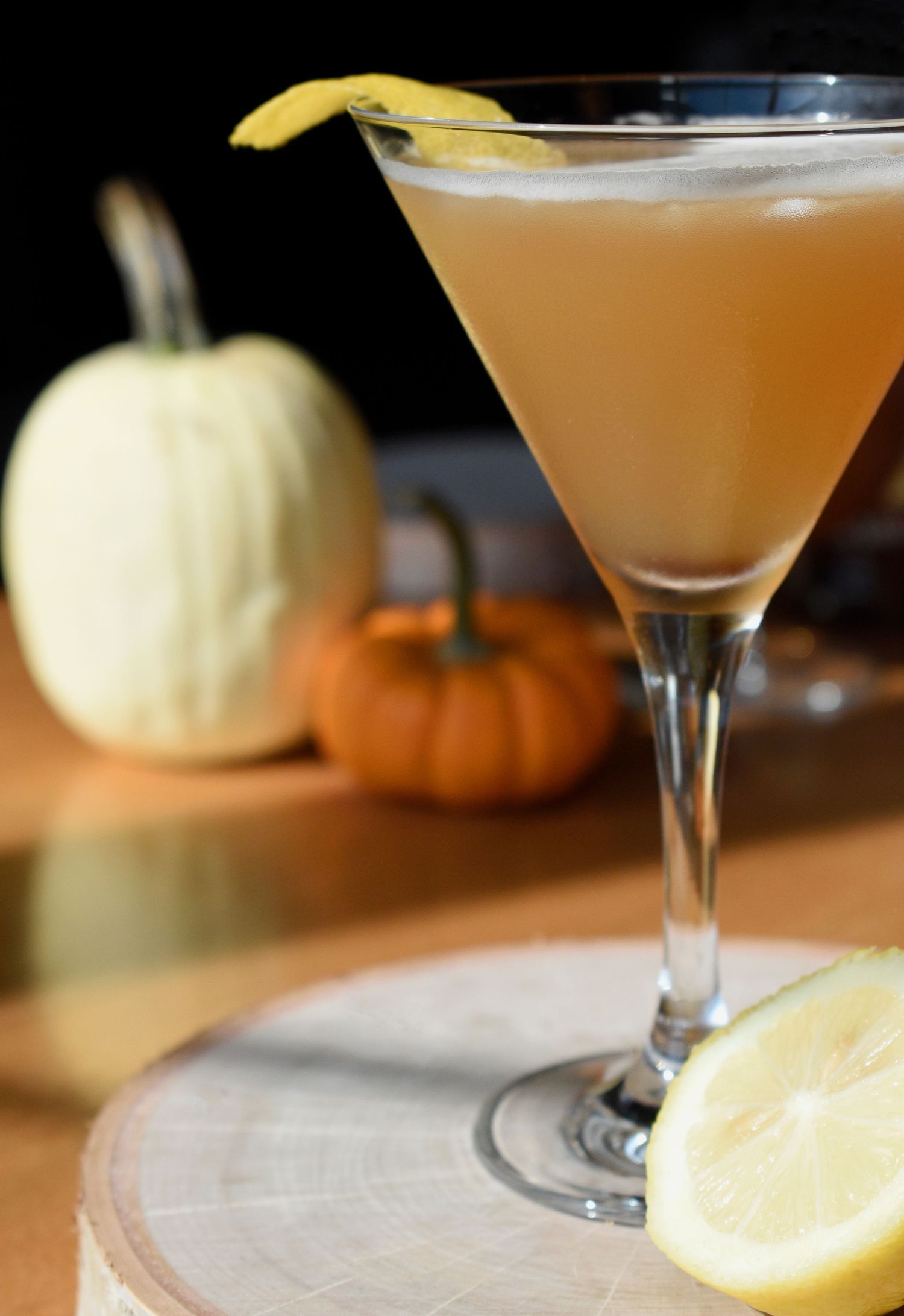 A golden amber cocktail in a martini glass with lemon twist garnish, displayed beside decorative mini pumpkins.