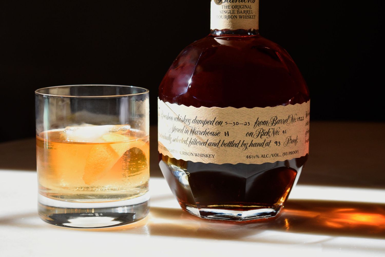 Premium bourbon whiskey bottle beside a rocks glass with ice on a sunlit surface.