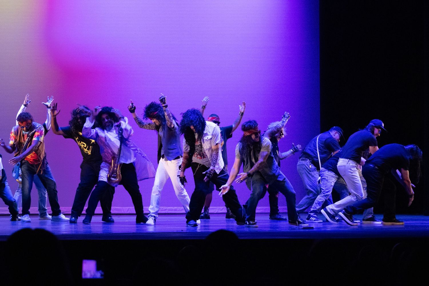 A large group performs an energetic hip-hop dance routine under colorful stage lights.