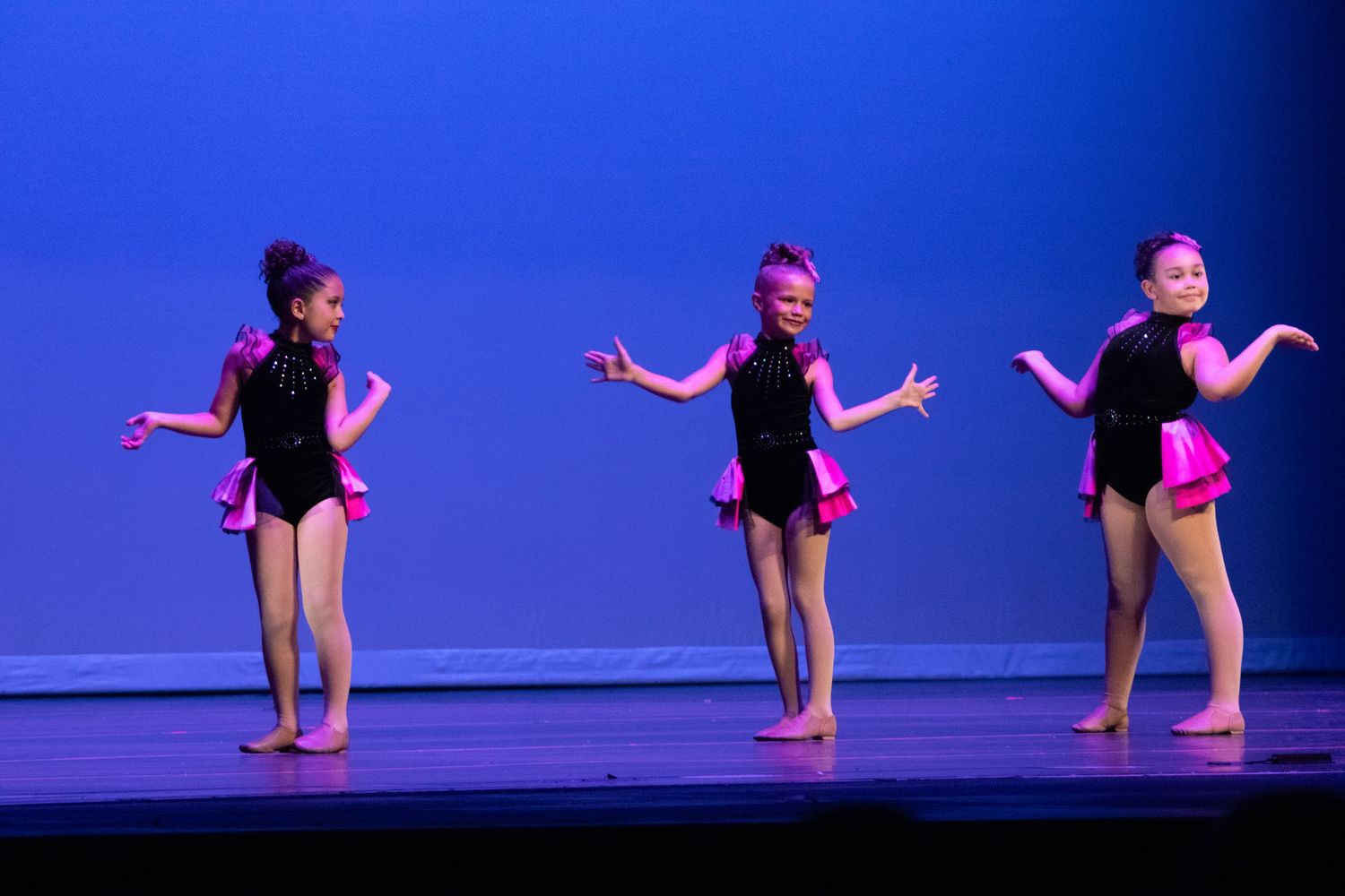Three young girls perform a jazz dance routine on stage wearing black and pink costumes under colorful stage lighting.