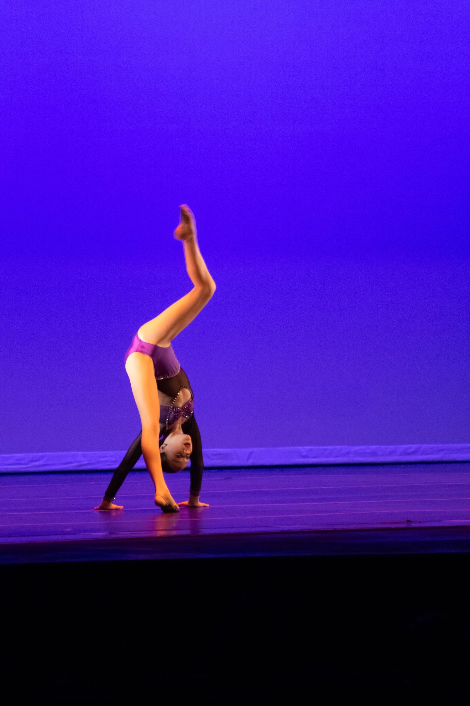 A dancer performs a handstand with one leg raised on a stage lit by vibrant blue lighting.