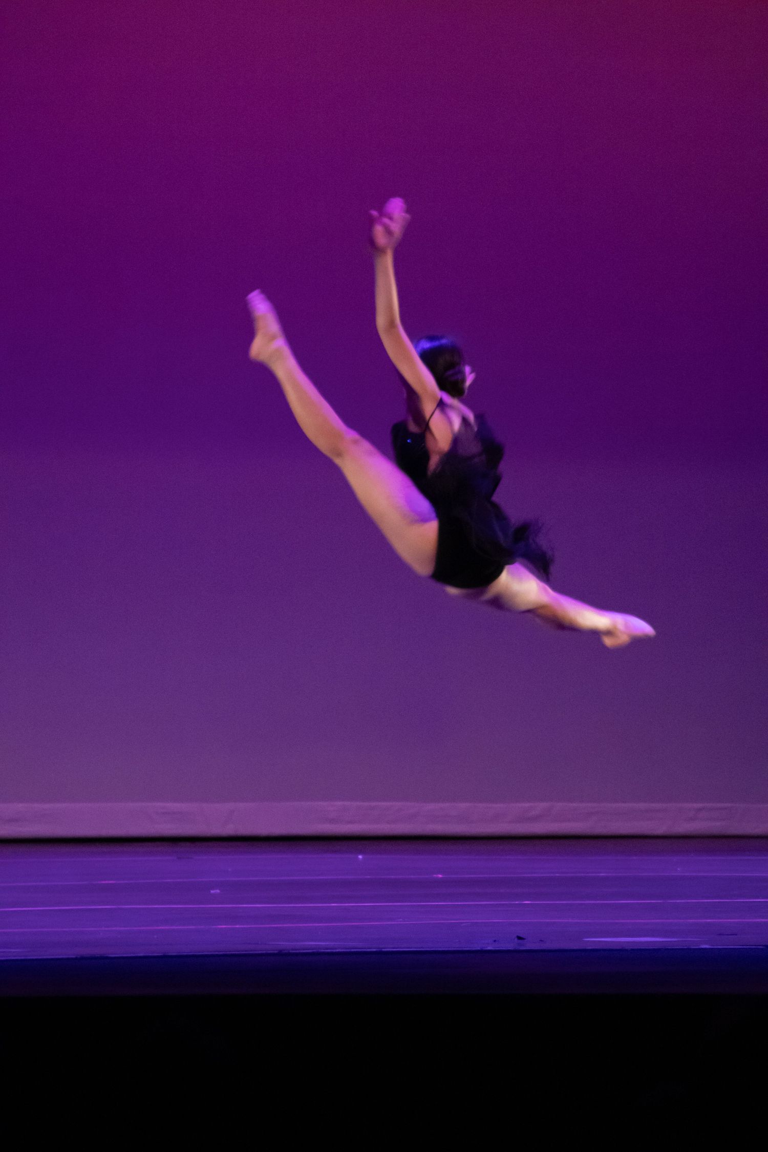 A dancer in a black leotard executes a stunning mid-air split leap on a purple-lit performance stage.