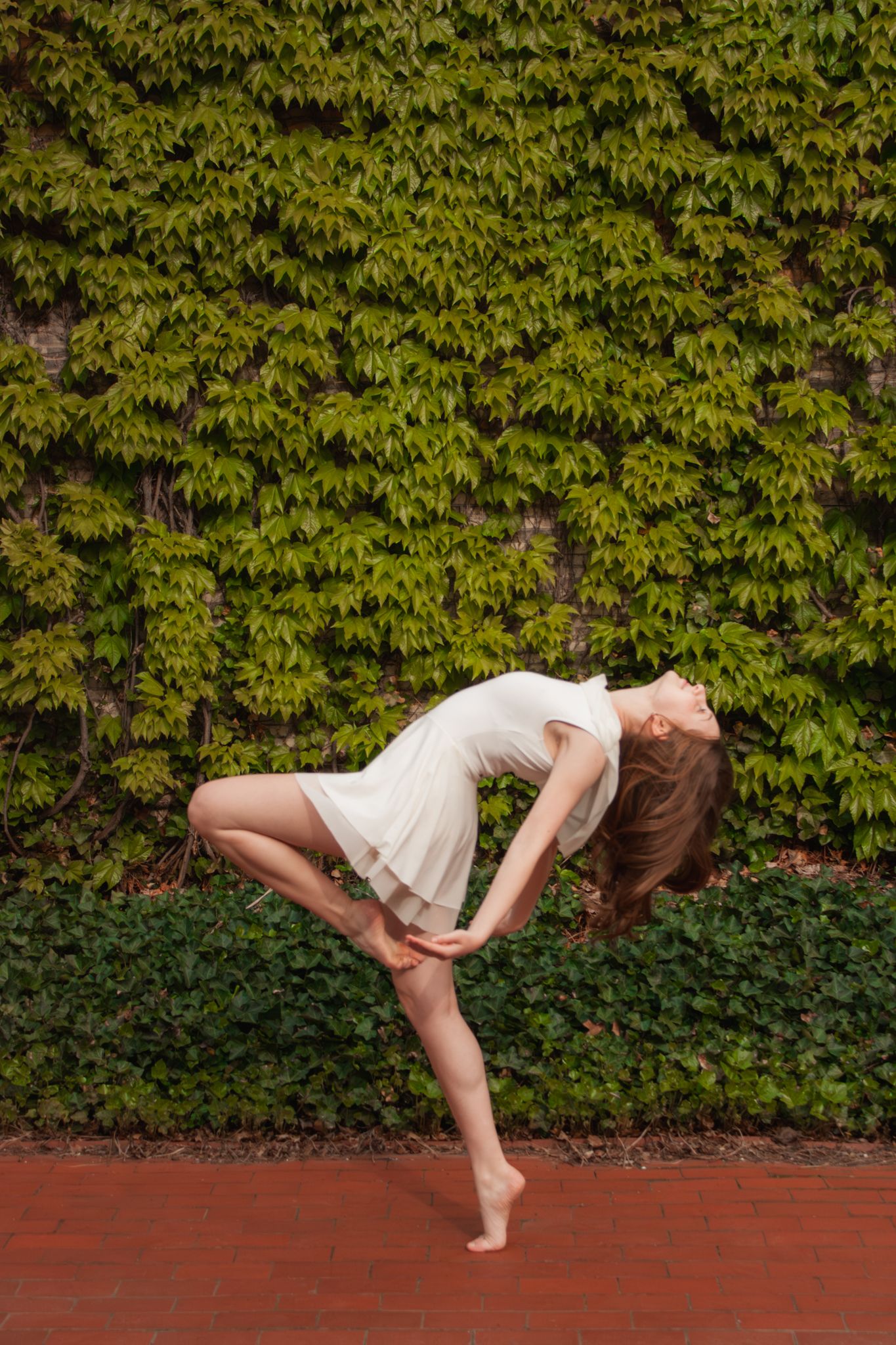 A barefoot dancer in a white dress performs a graceful backbend pose outdoors against a lush green ivy wall.
