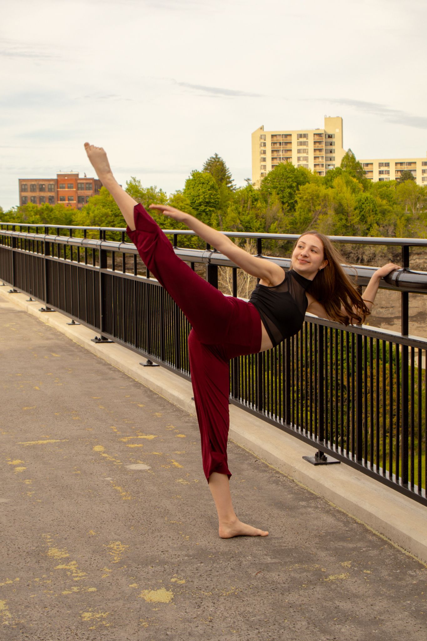Female dancer executes a high side kick on a bridge with urban skyline in the background.