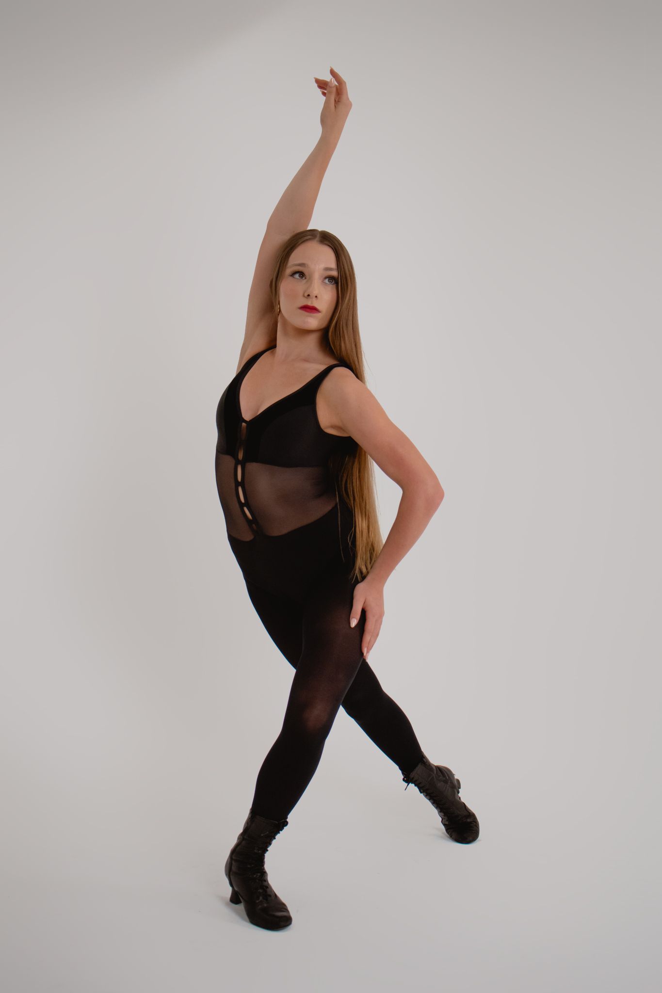 Female dancer in black bodysuit and boots striking a ballet-inspired pose in a white studio.