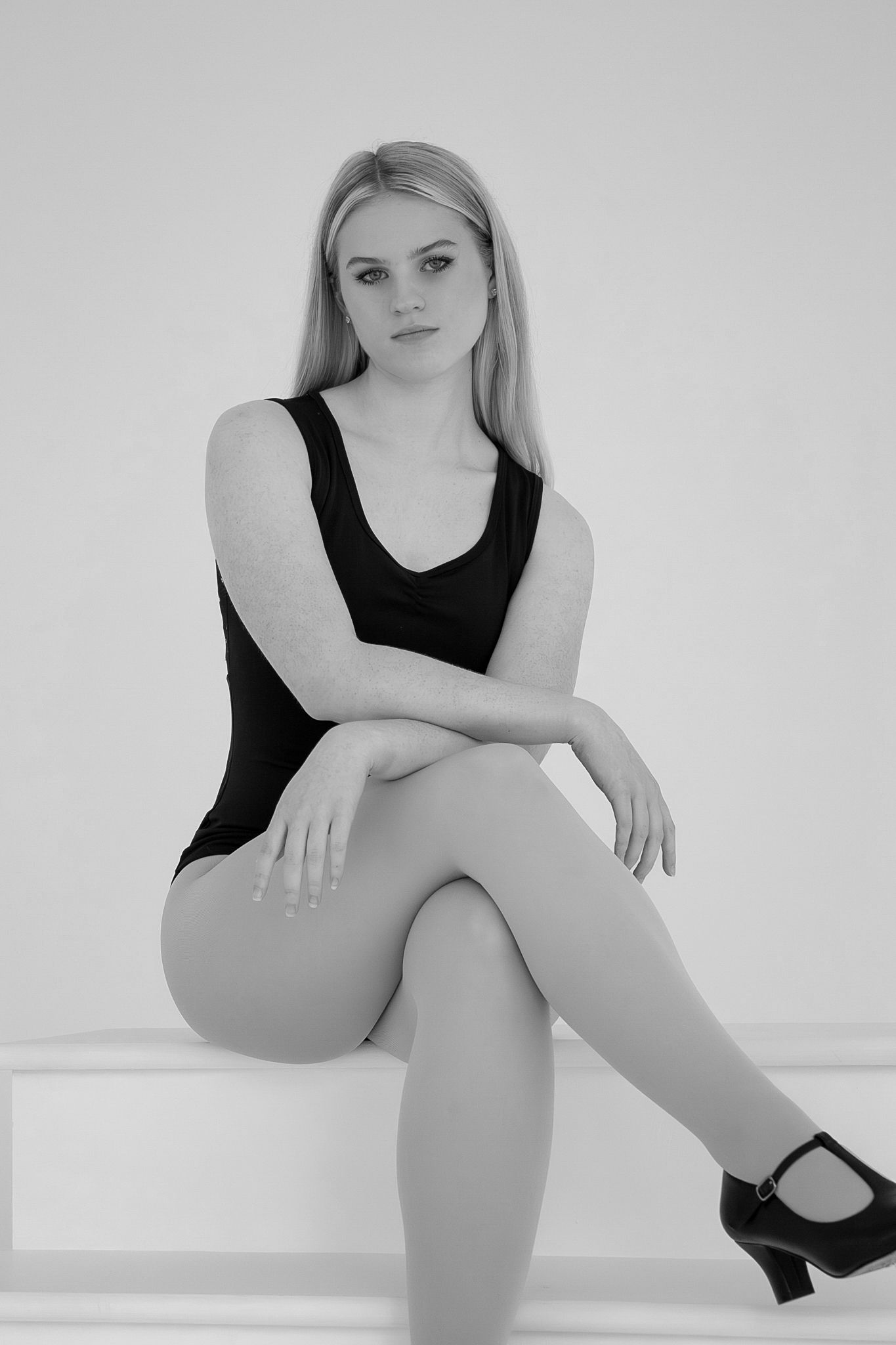 Black and white portrait of blonde dancer in black bodysuit seated on white platform.
