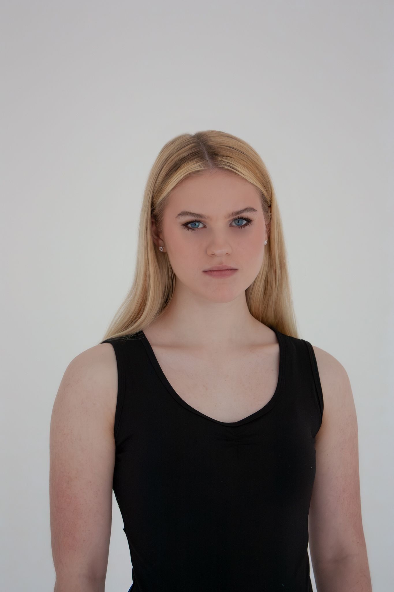 Young blonde woman with blue eyes wearing a black tank top poses against a white background.