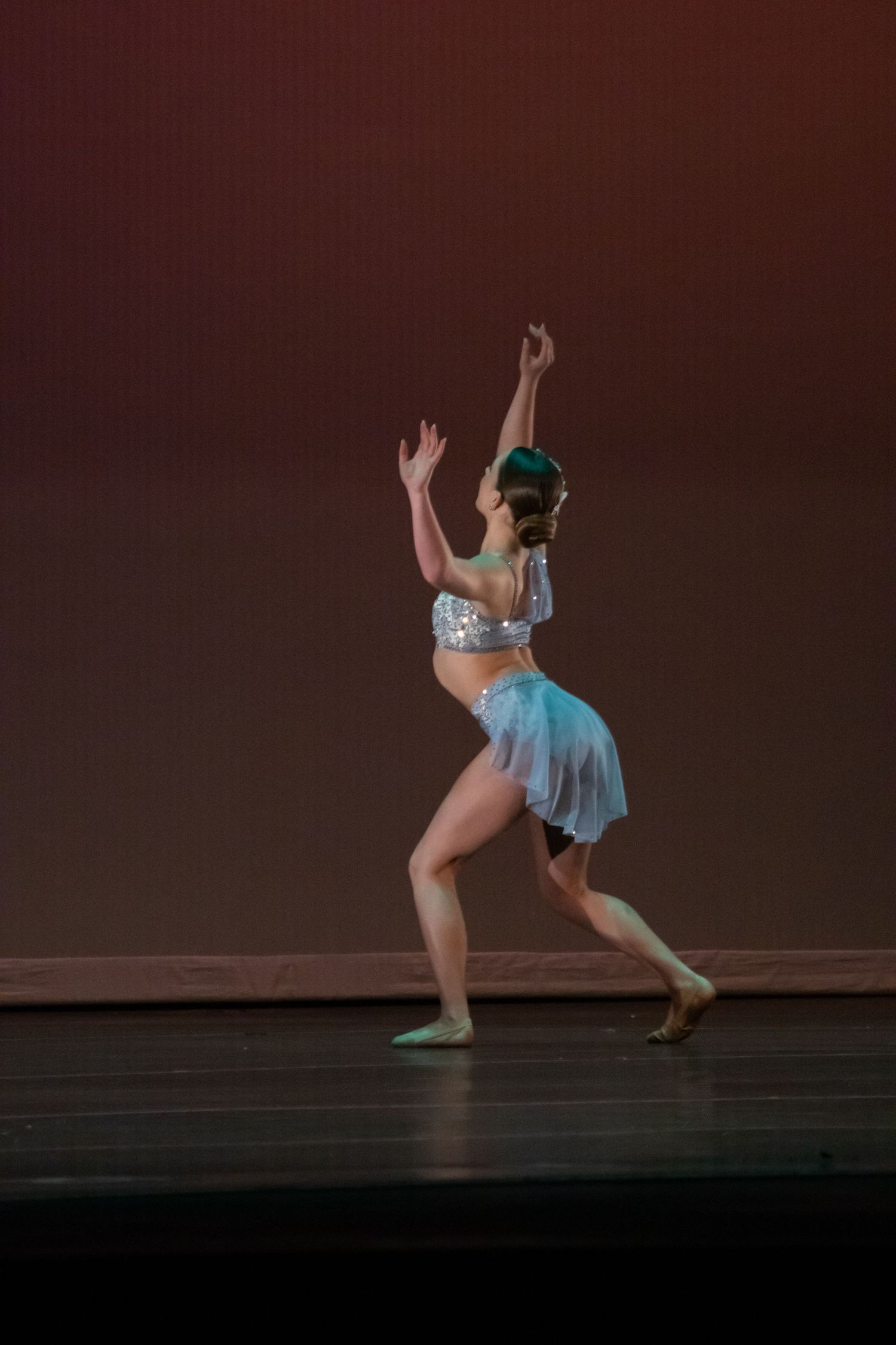 Female dancer in silver and white costume reaches upward on a warmly lit stage.
