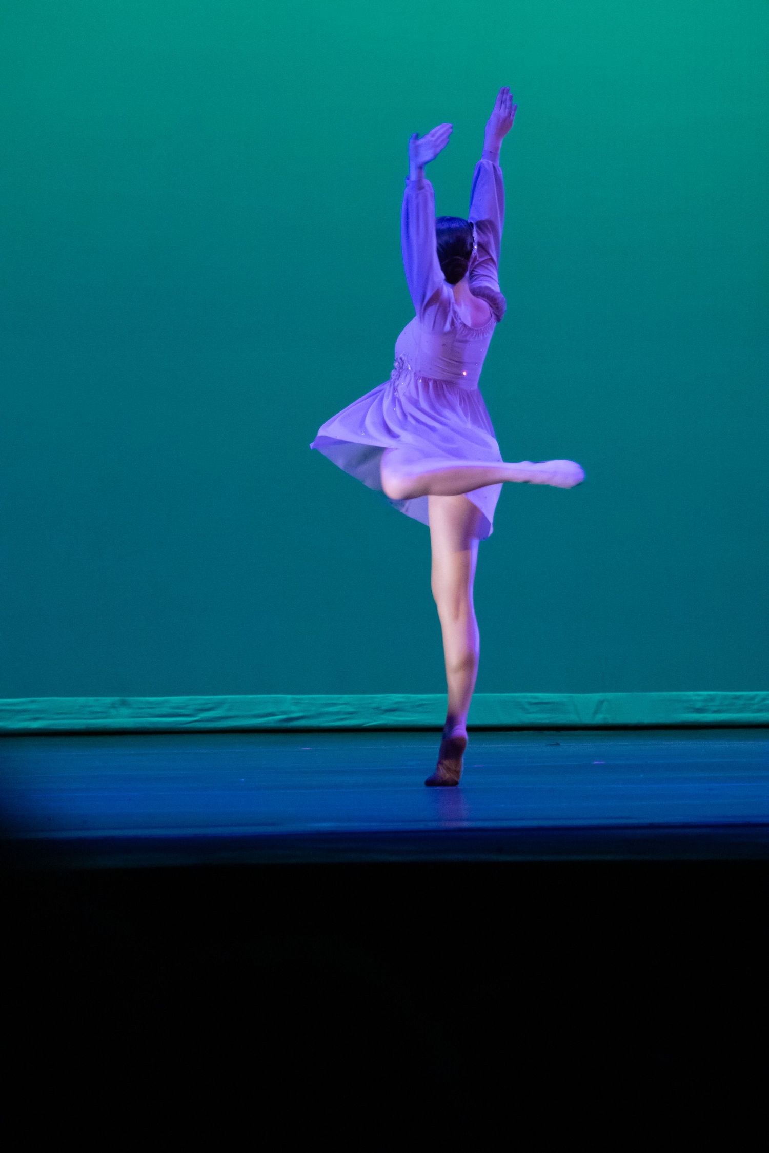A ballet dancer in a purple dress elegantly balances on one leg with arms raised on a teal-lit stage.