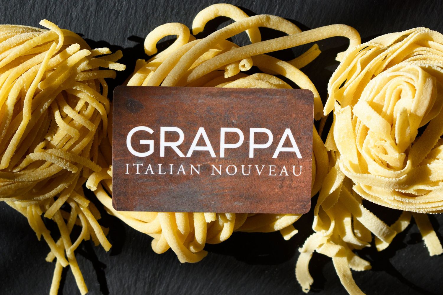 Grappa Italian Nouveau gift card displayed among fresh uncooked tagliatelle and spaghetti pasta nests.