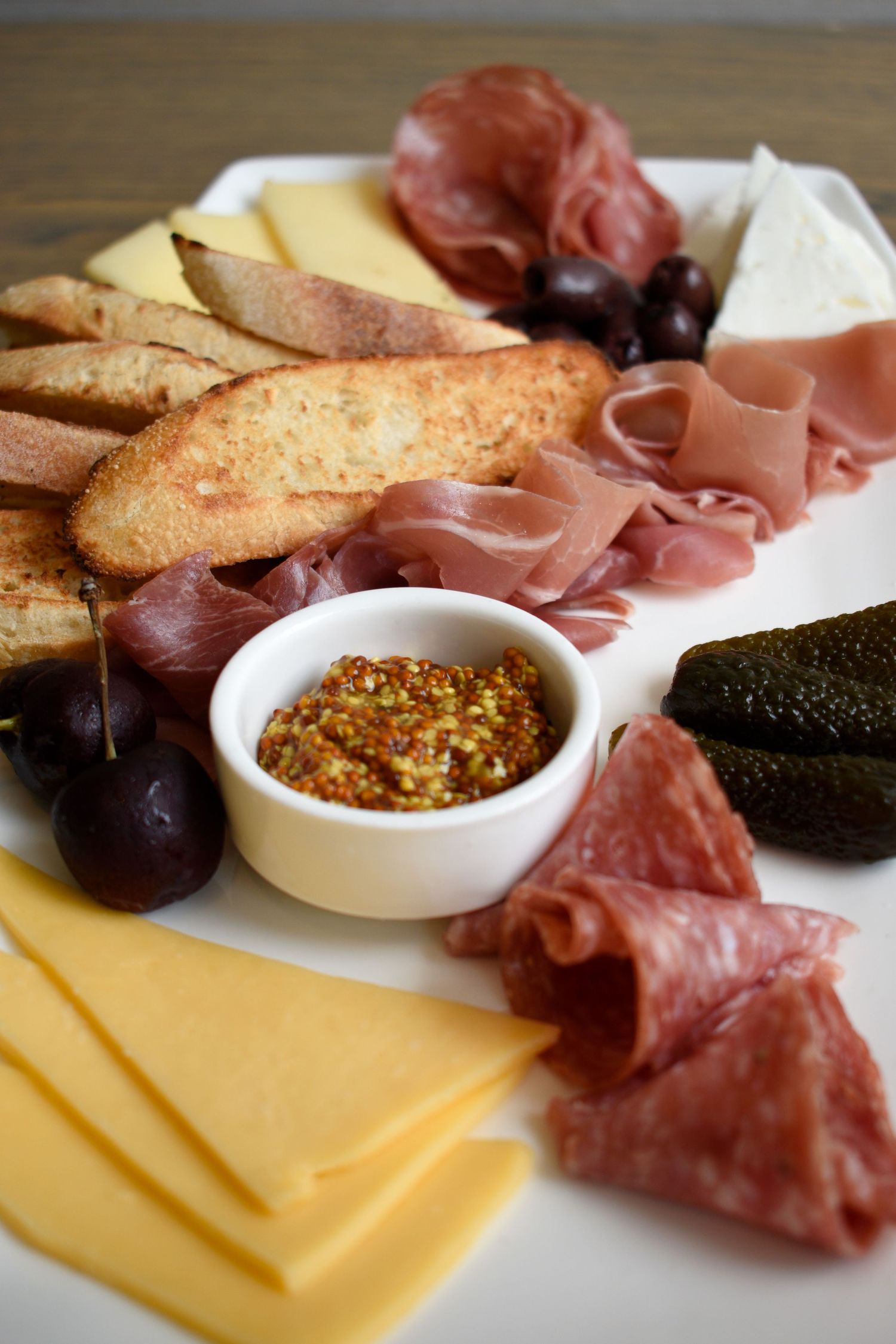Charcuterie platter with assorted cured meats, yellow and white cheese, olives, toasted bread, and mustard.