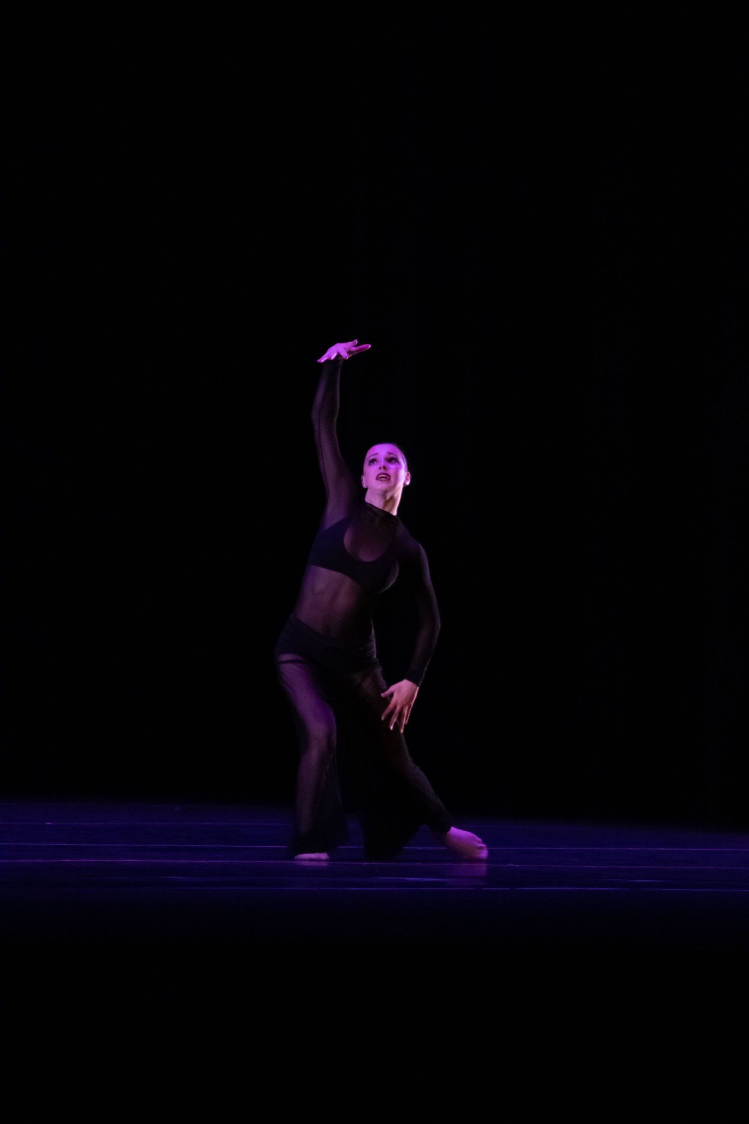 A dancer in black performs on a dark stage under purple lighting with one arm raised gracefully overhead.