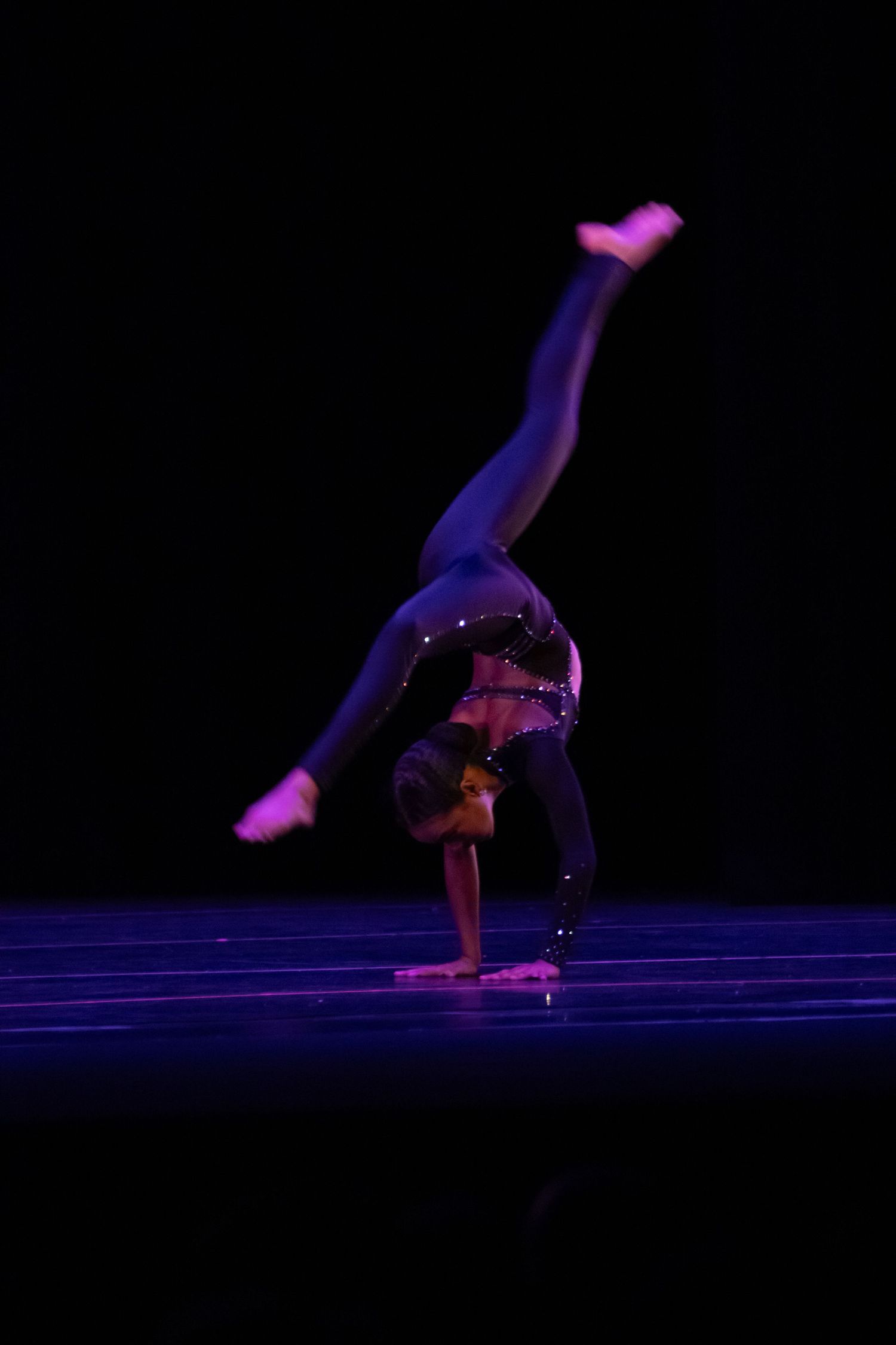A dancer executes a one-handed cartwheel in a black costume on a dark stage.