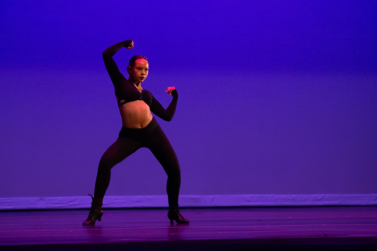 Pregnant dancer performs on stage in black outfit under purple lighting, striking a powerful pose with arms raised.