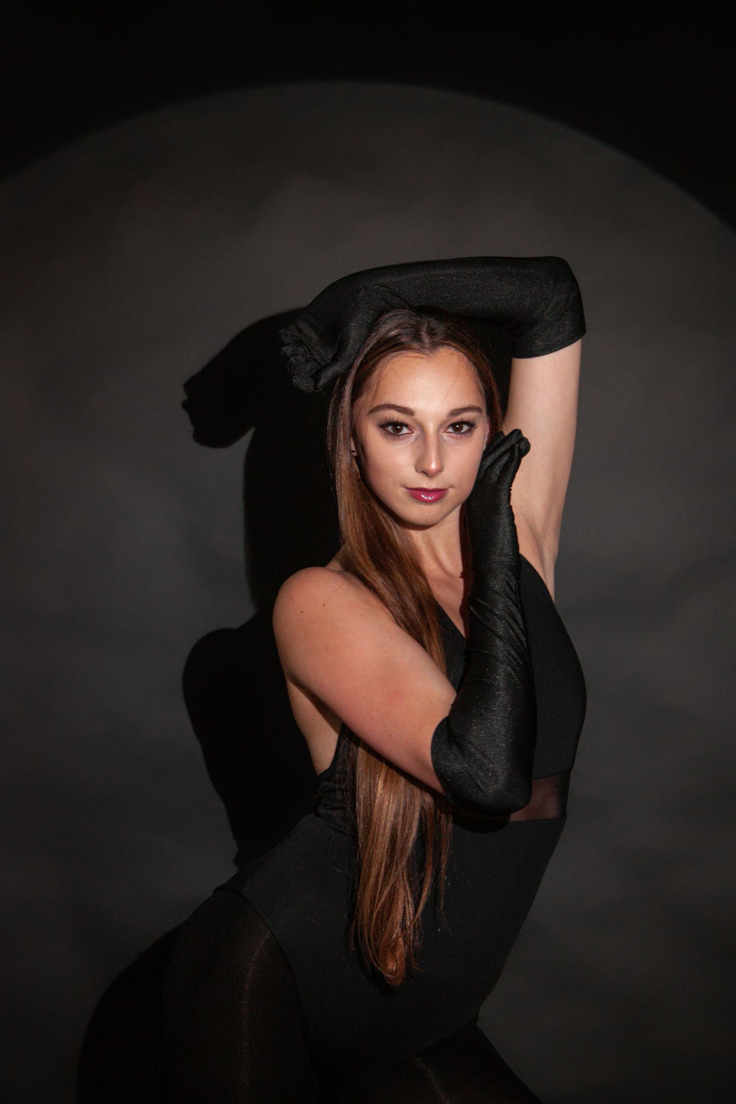 Young woman in black gloves and outfit poses gracefully against dark studio backdrop with dramatic lighting.