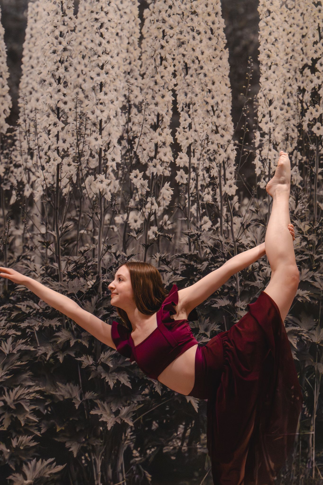 Elegant dancer performs a high leg extension in burgundy outfit against a floral botanical backdrop.