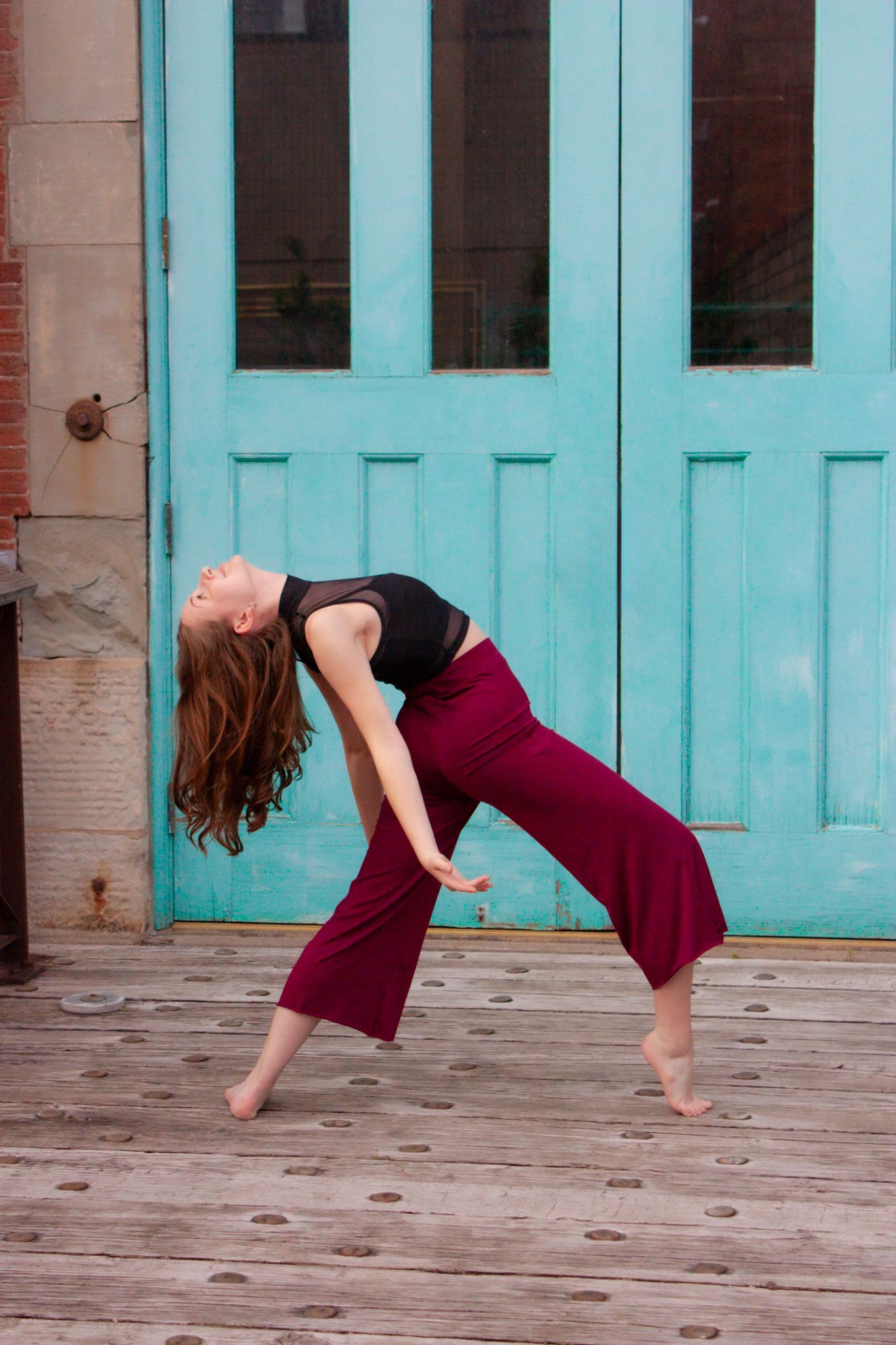 Young female dancer in dramatic backbend with flowing hair, wearing burgundy pants and black crop top outdoors.