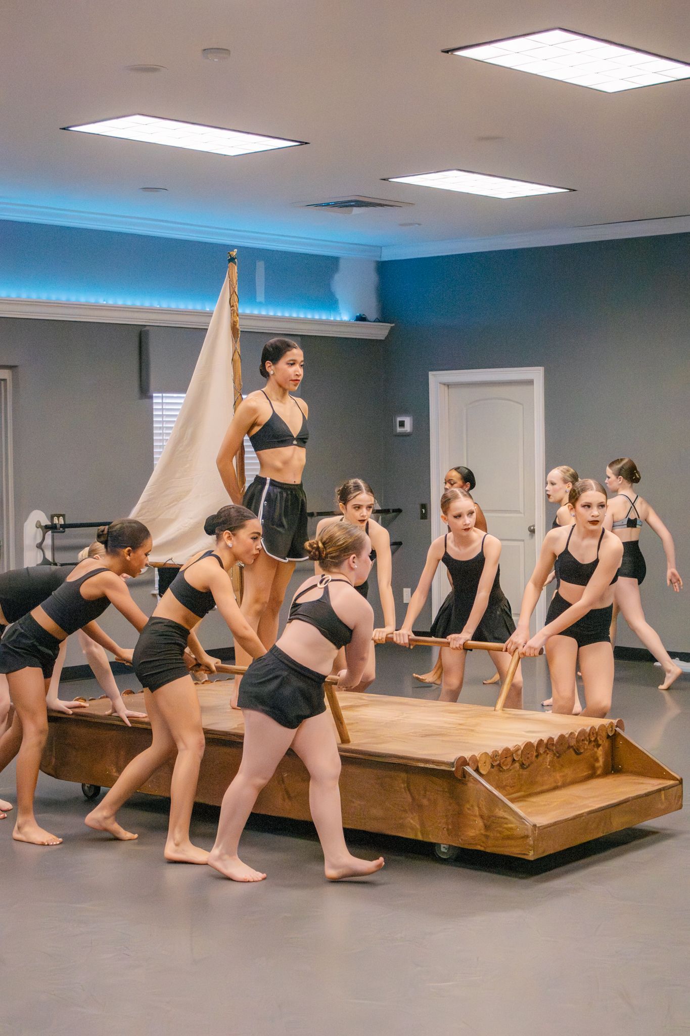 Dance students in black costumes rehearsing with wooden boat prop and sail in dance studio.