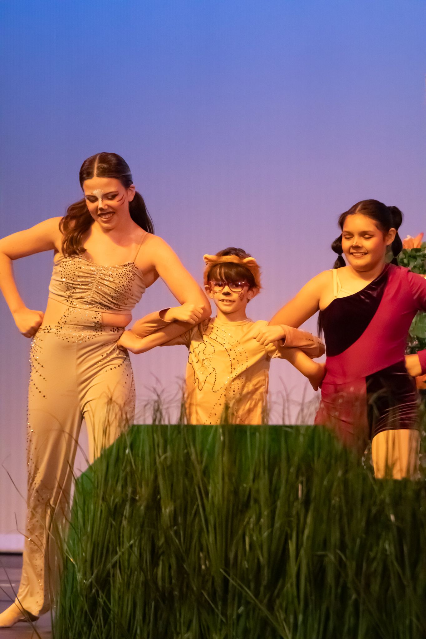 Three performers in a dance recital scene with grass props under warm stage lighting.