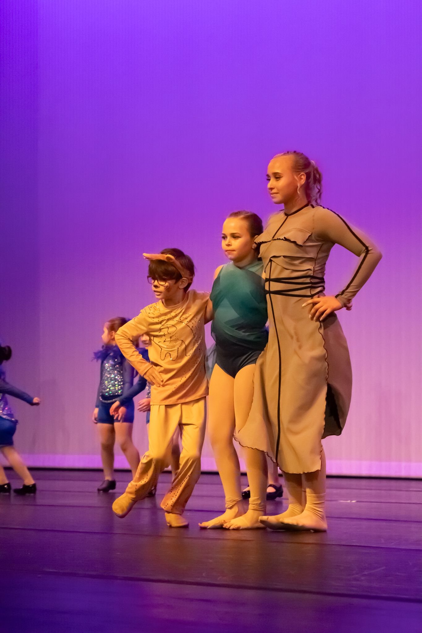 Three young dancers perform on stage in colorful costumes under purple lighting.