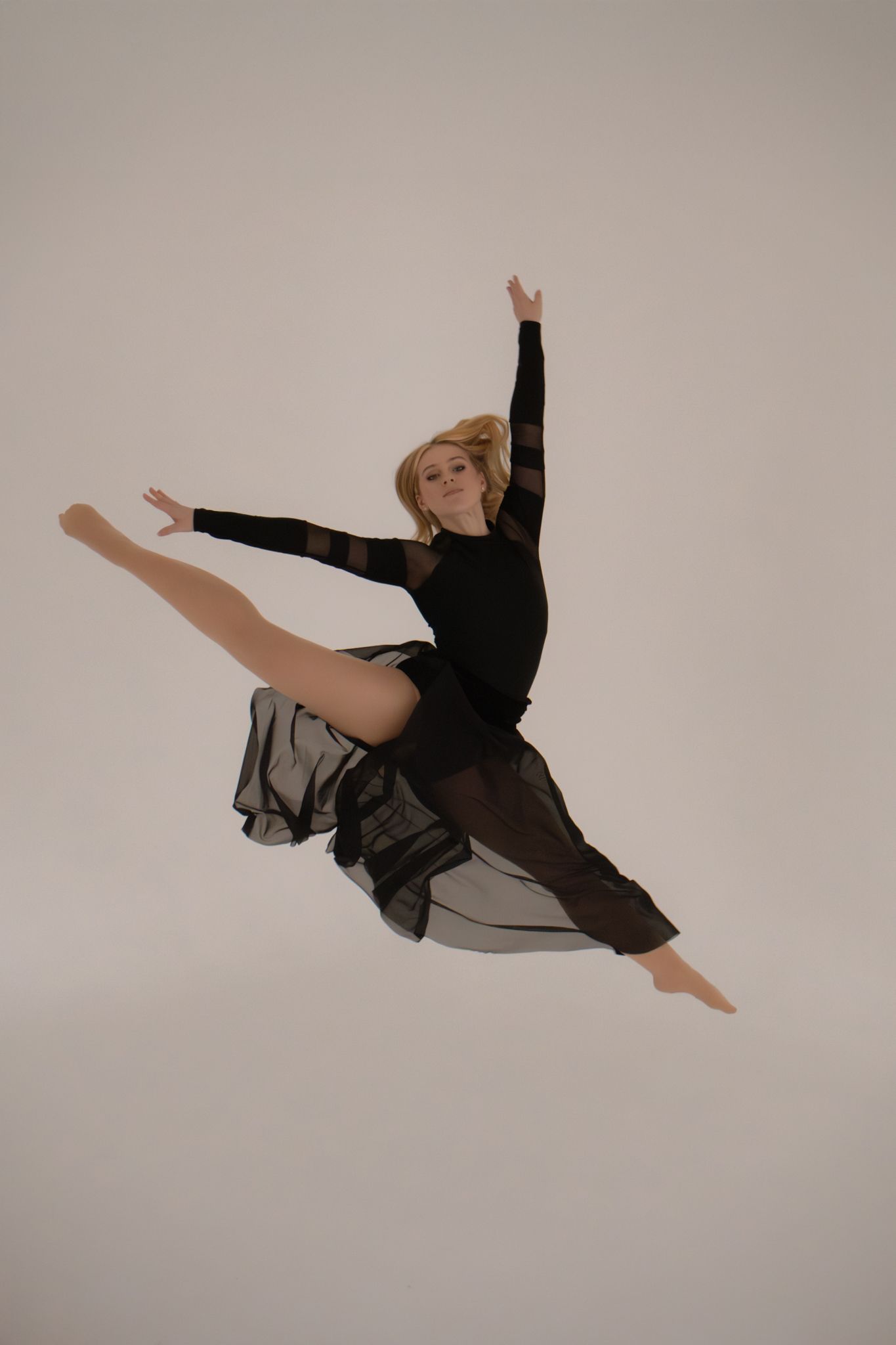 Blonde dancer leaps mid-air in black bodysuit and flowing sheer skirt against white background.