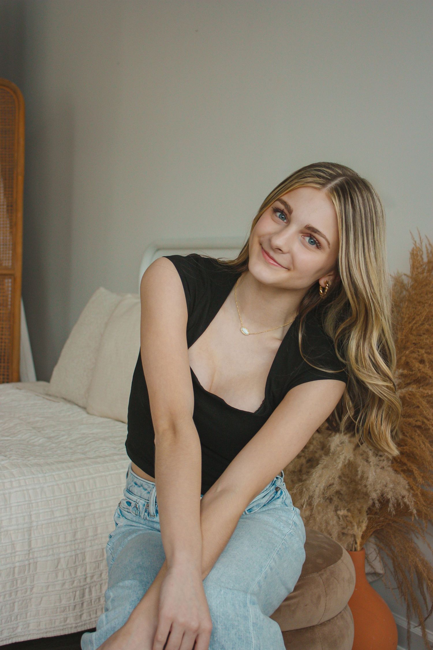 Young woman with blonde hair wearing black top and jeans, sitting and smiling in a cozy bedroom setting.
