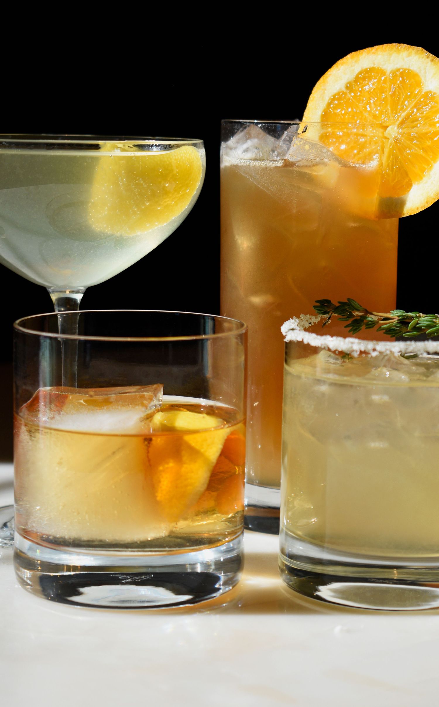 Four elegant cocktails garnished with citrus fruits and herbs arranged on a white surface against black background.