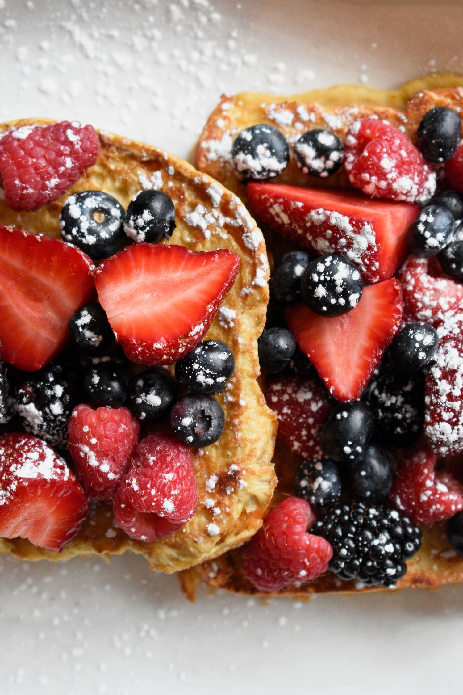 French toast topped with fresh strawberries, blueberries, raspberries, and powdered sugar on a white plate.