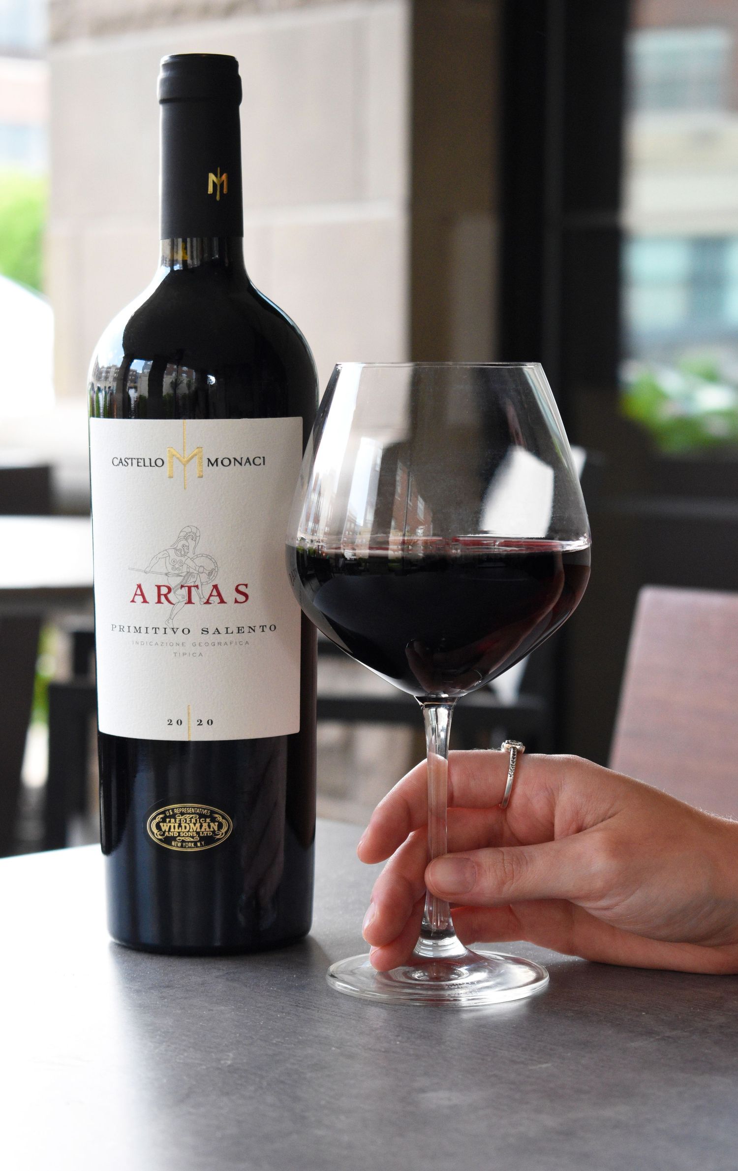 Hand holding a glass of Castello Monaci Artas Primitivo Salento red wine next to the bottle on a table.