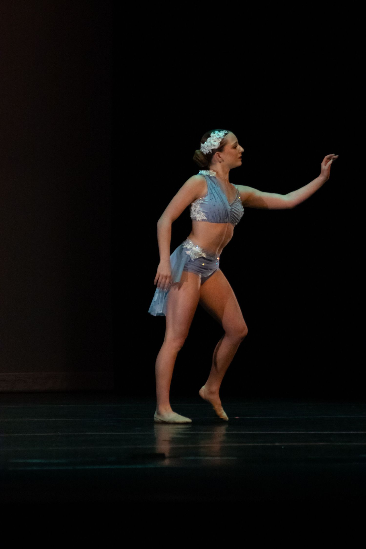 A female dancer in a blue costume performs a lyrical solo on a darkly lit stage.