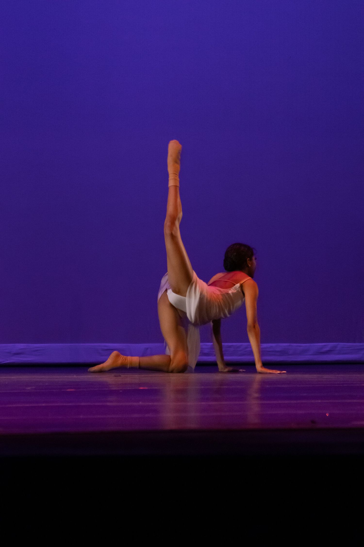 A gymnast dancer kneels on stage extending one leg high while wearing a red and white costume on a purple-lit stage.