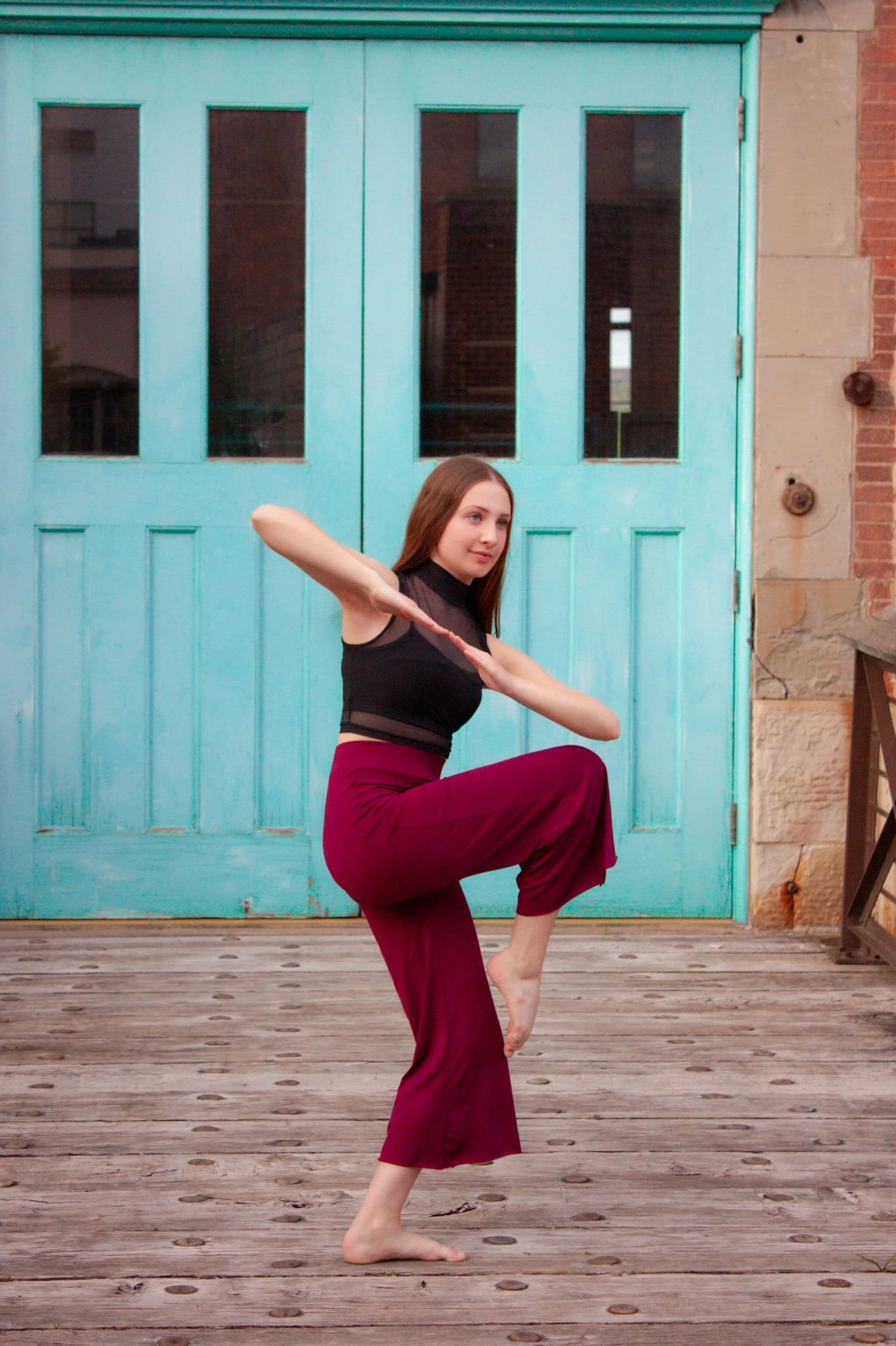 Dancer balancing on one leg in red pants and black top before teal wooden doors.