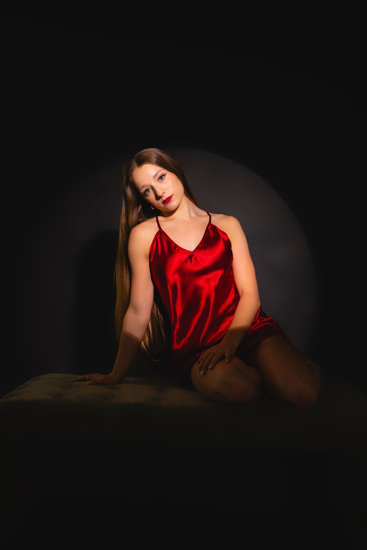 Young woman in red satin slip dress sits gracefully under spotlight on dark background.