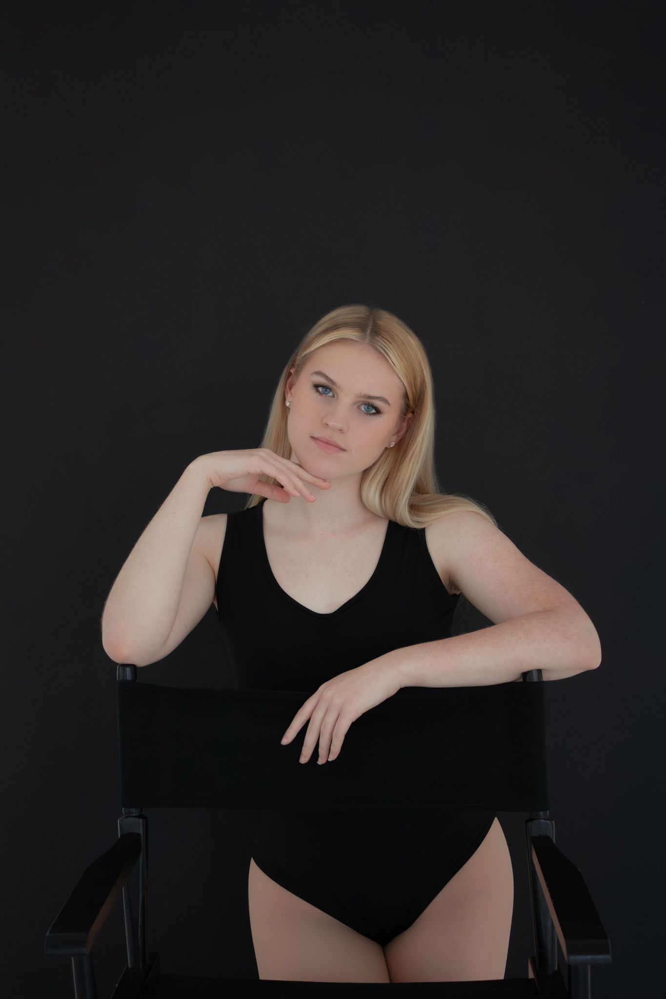 Blonde woman in black bodysuit posing on chair against dark background.
