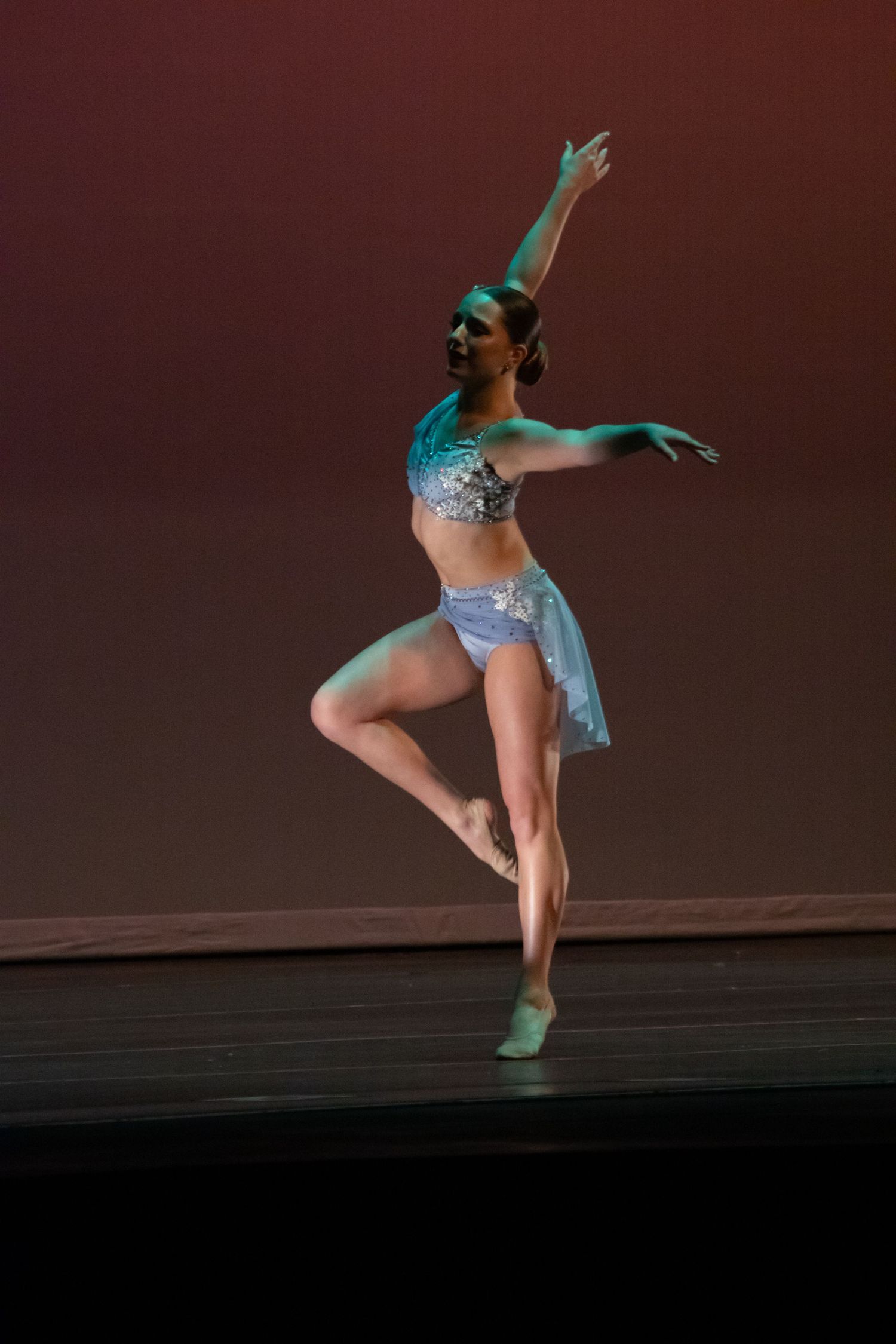 Dancer in teal and silver costume balancing on pointe under warm stage lighting.