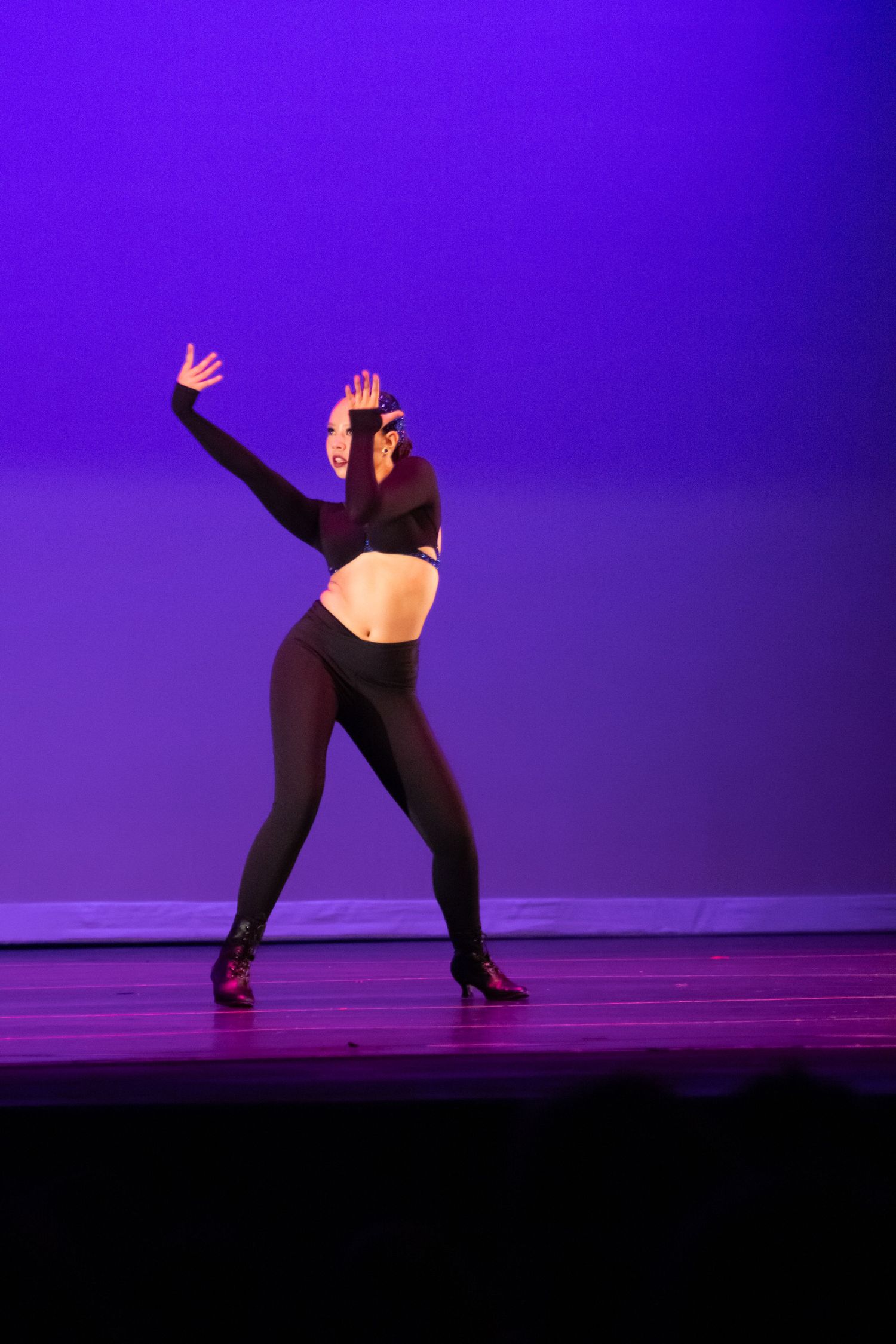 Dancer in black crop top and tights striking a bold pose on a purple-lit stage.