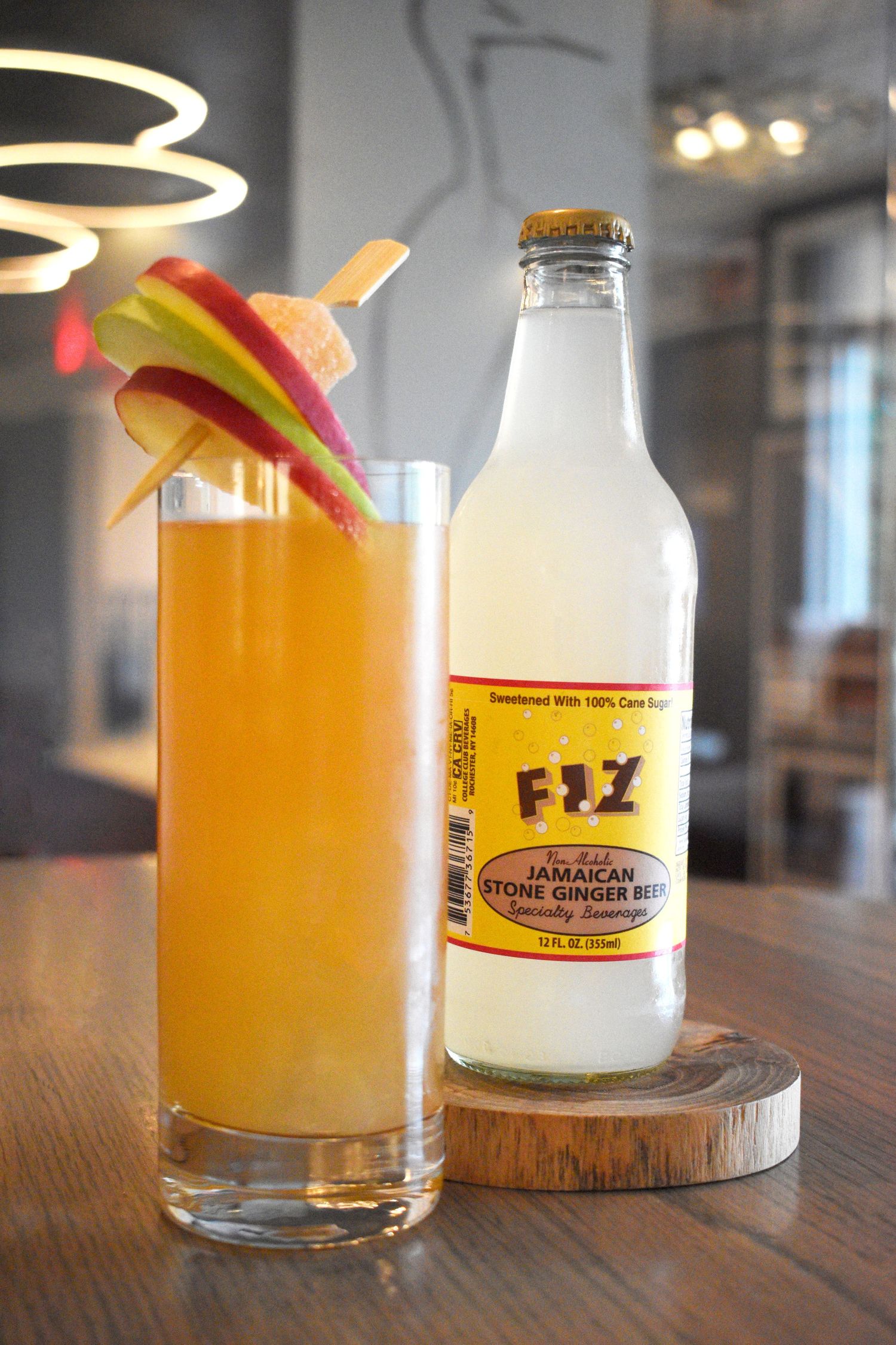 Golden ginger beer cocktail with apple garnish beside a Fizz Jamaican Stone Ginger Beer bottle on a wooden coaster.