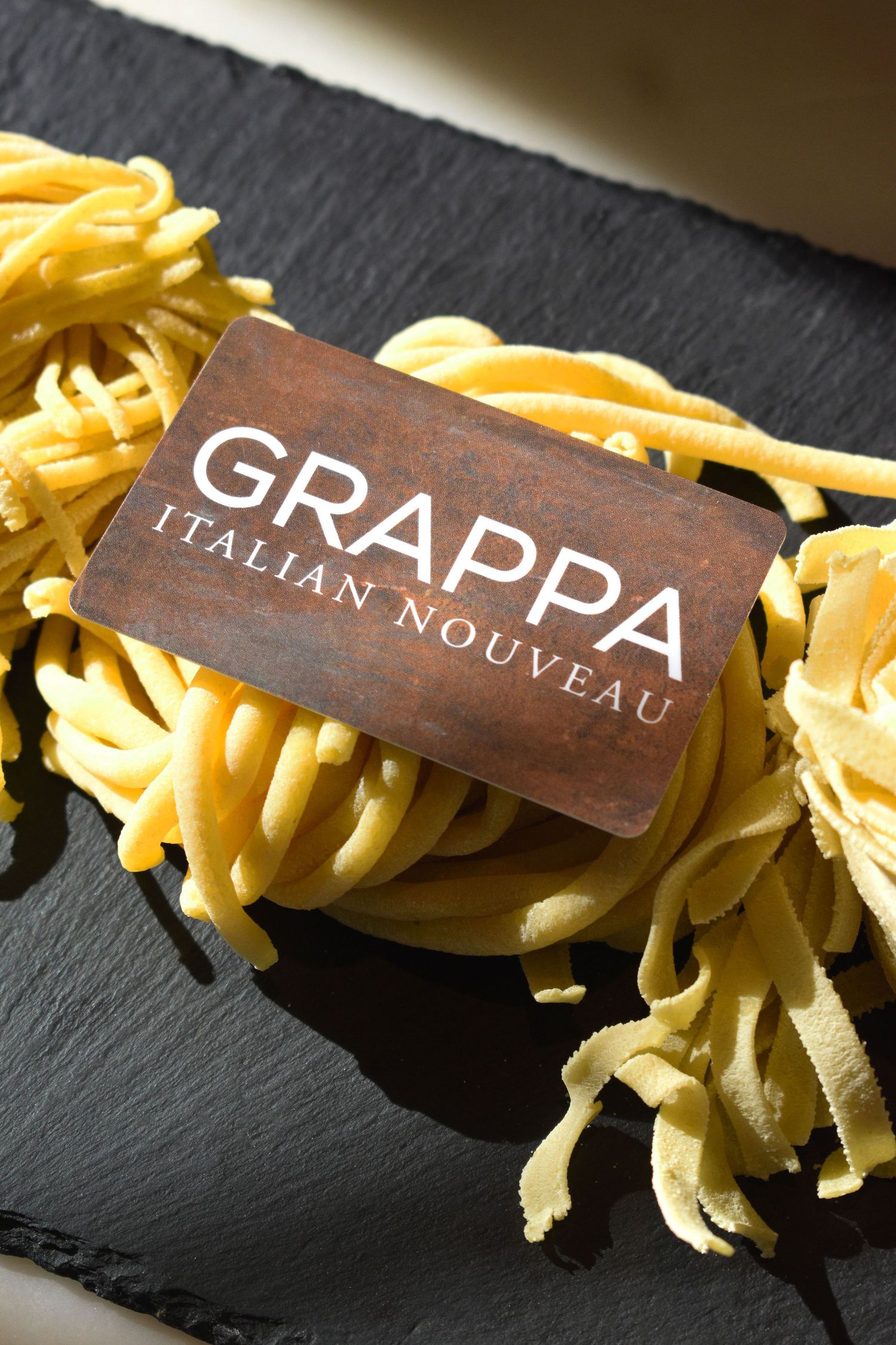 Grappa Italian Nouveau gift card placed on fresh pasta nests on dark slate board.