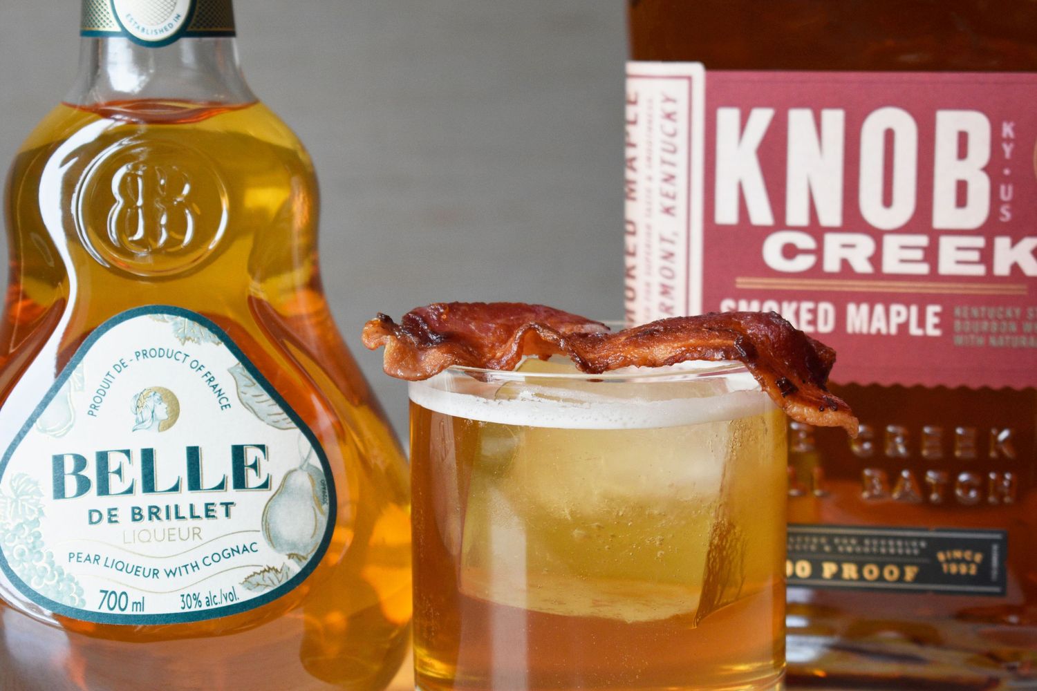 Knob Creek smoked maple whiskey cocktail garnished with crispy bacon beside Belle de Brillet liqueur bottle.