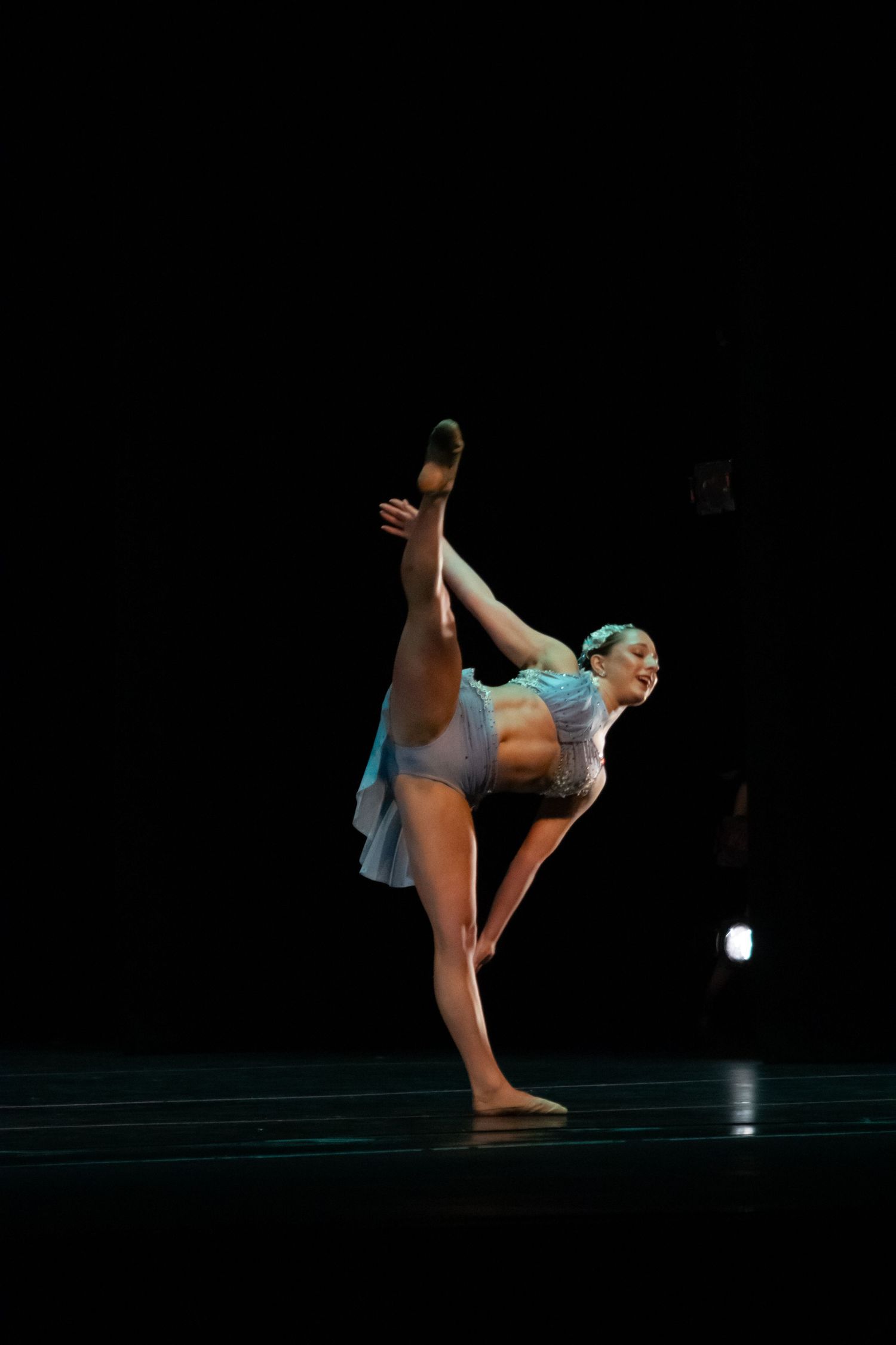 A dancer performs a high leg extension in a blue costume under dramatic black stage lighting.
