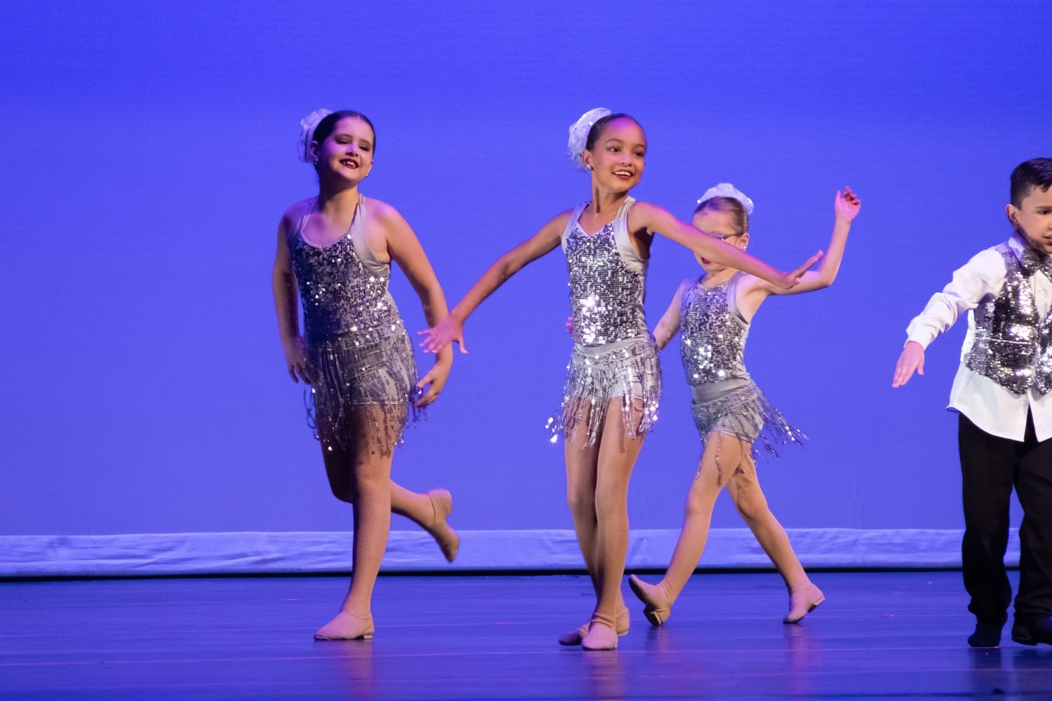 Three girls in silver sequined dresses and a boy in white jacket dance together on stage.