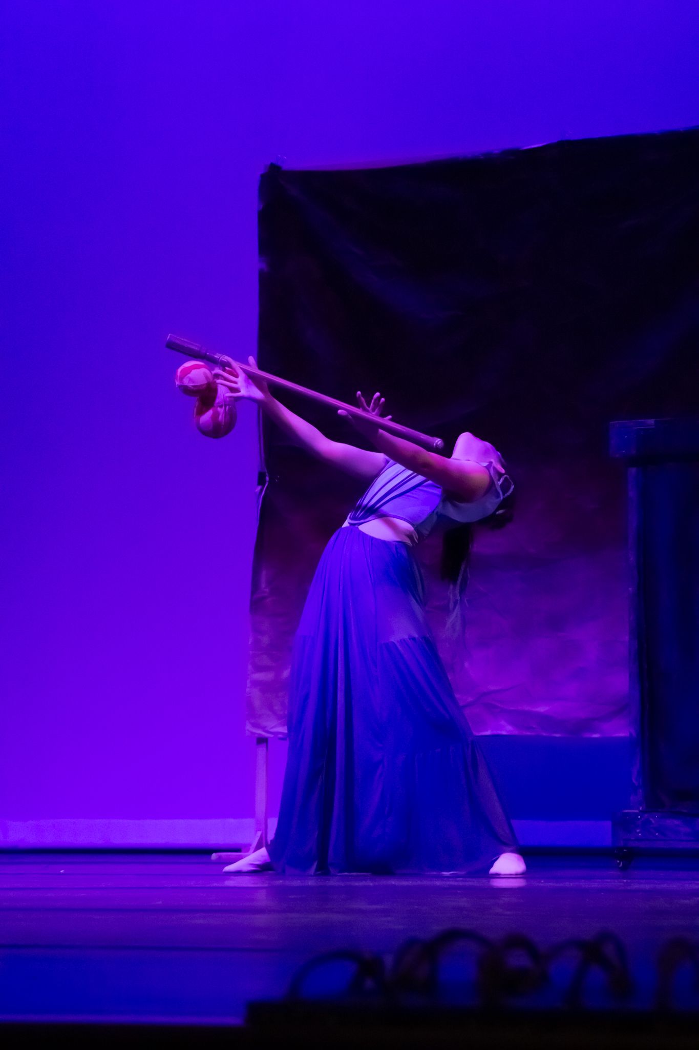 Solo dancer in blue skirt dramatically poses with a staff under purple stage lighting.