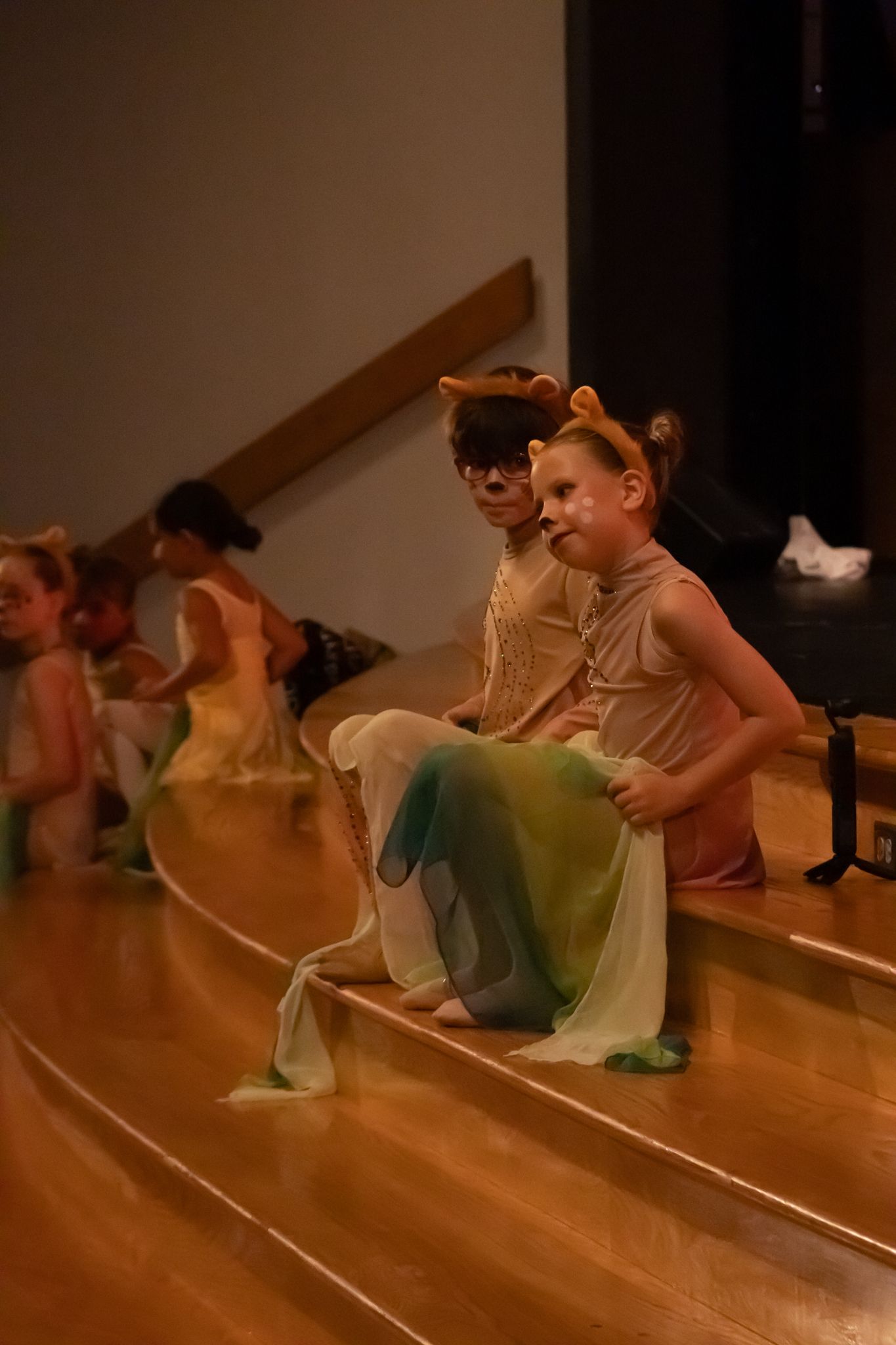 Young children in lion costumes with green scarves sit on stage steps during a dance recital performance.