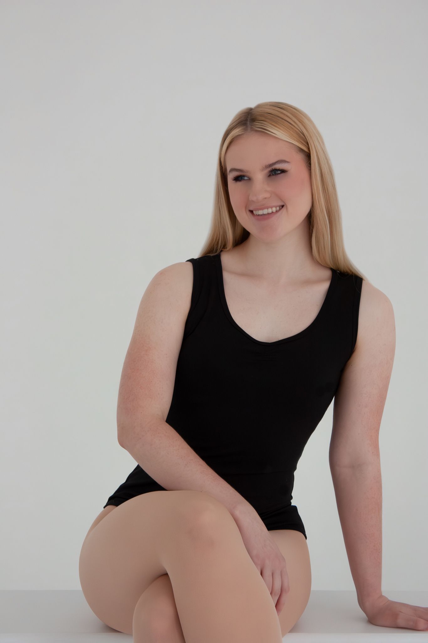 Smiling blonde woman in black tank leotard sitting gracefully against white background.