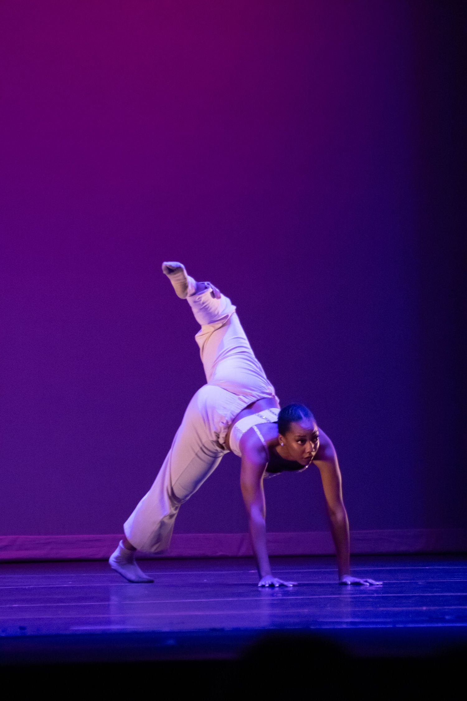A dancer in white performs an acrobatic one-handed pose with leg raised on a purple-lit stage.